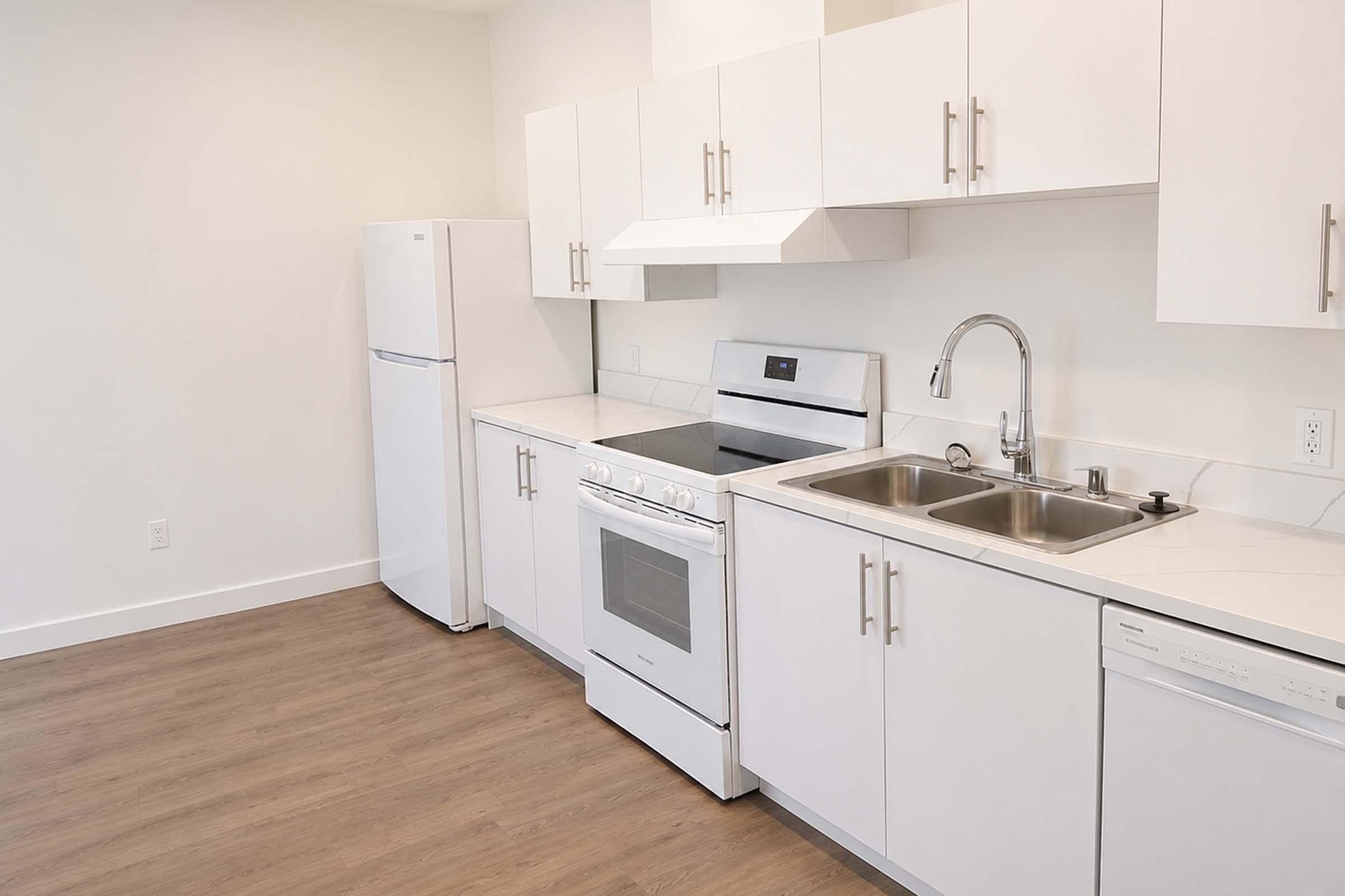 A modern kitchen featuring white cabinetry, a stainless steel stove and oven, a double sink, and a refrigerator. The countertop is light-colored, and the flooring is a warm-toned wood. The walls are painted white, creating a clean and bright atmosphere.