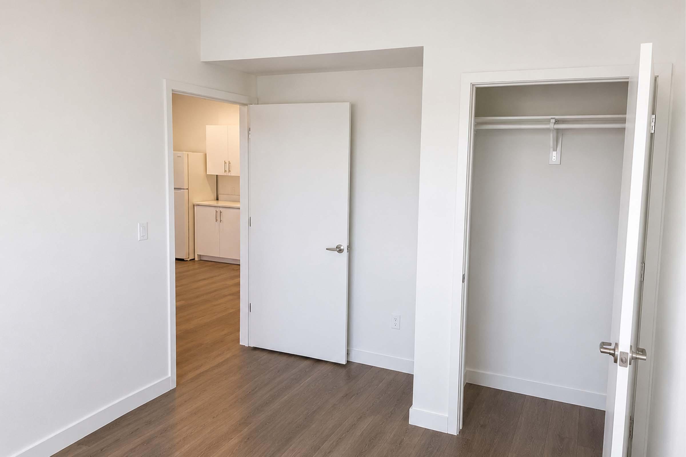 A minimalist room featuring light-colored walls and flooring. There's a door leading to a kitchen area partially visible in the background. To the right, an open closet with a hanging rod is displayed. Natural light brightens the space, giving it an airy feel.
