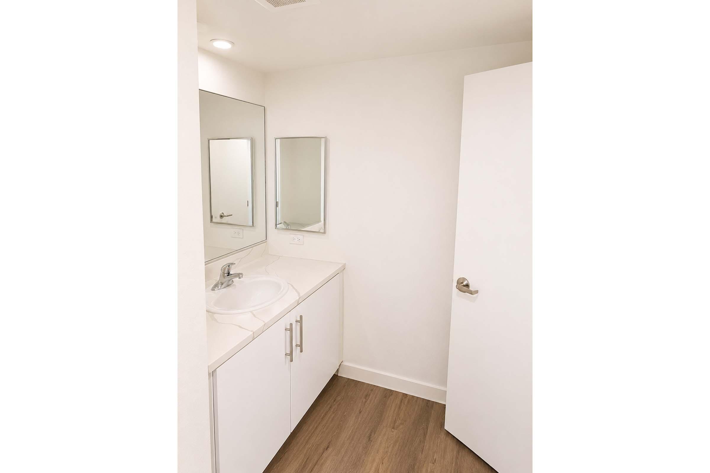 A modern bathroom featuring a white vanity with a sink, two mirrors above the sink, and a door leading to another area. The walls are light-colored, and the flooring is wooden, contributing to a sleek and clean design. The overall ambiance is bright and minimalist.