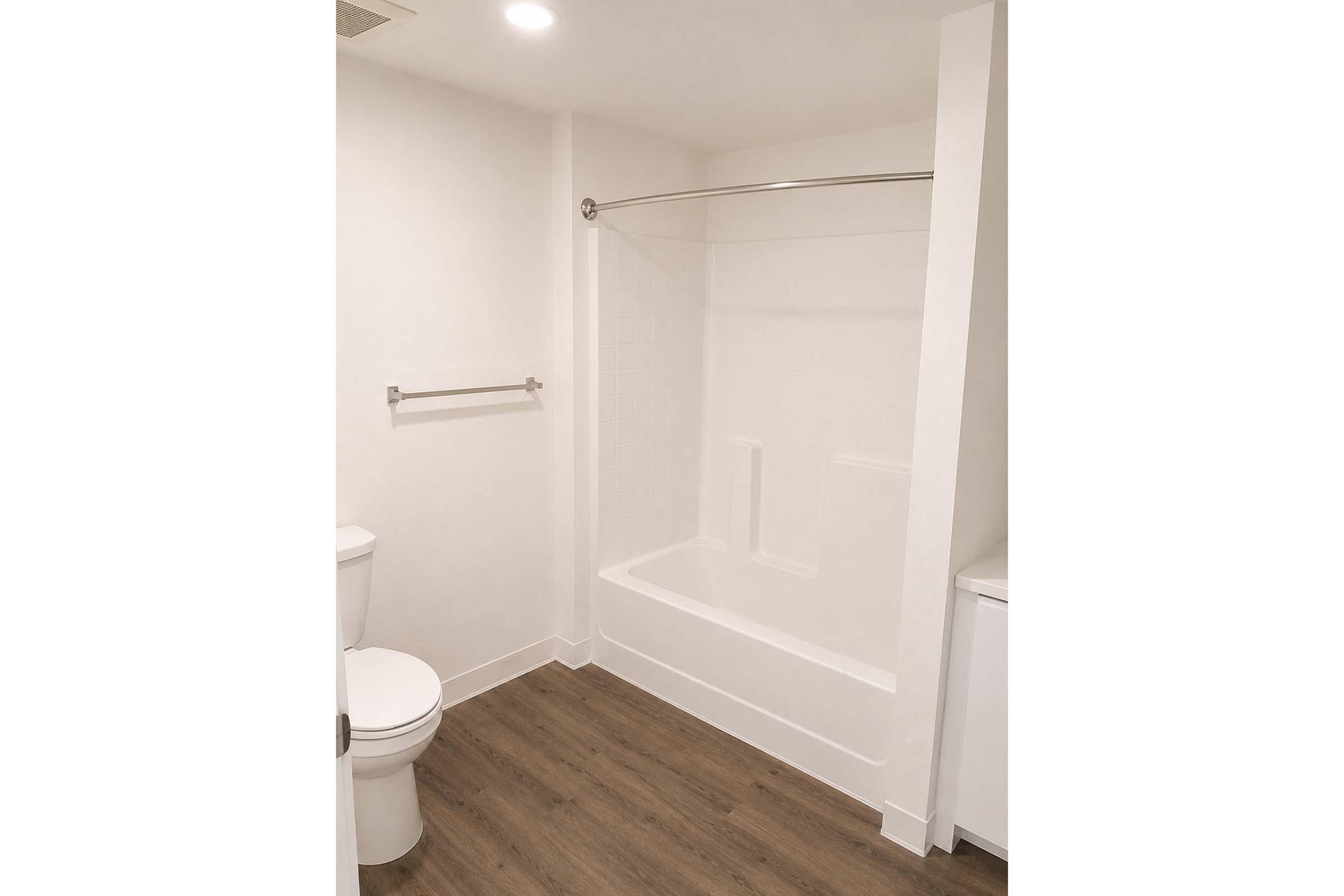 A clean, modern bathroom featuring a white bathtub with a shower rod, a toilet, and a towel bar. The walls are painted white, and the flooring is a light wood laminate. The overall design is simple and contemporary, providing a bright and spacious feel.