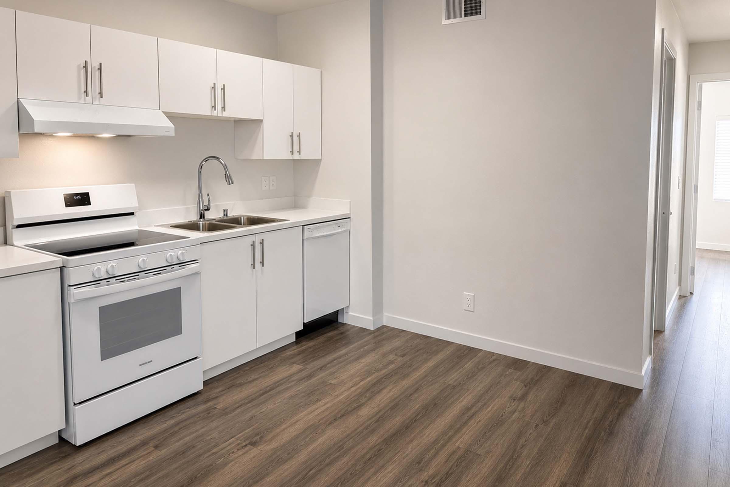 A modern kitchen featuring white cabinetry, a stainless steel sink, and a gas stove. A dishwasher is also present. The space has light-colored walls and laminate flooring, with a doorway leading to another area. Natural light is visible from the adjacent room.