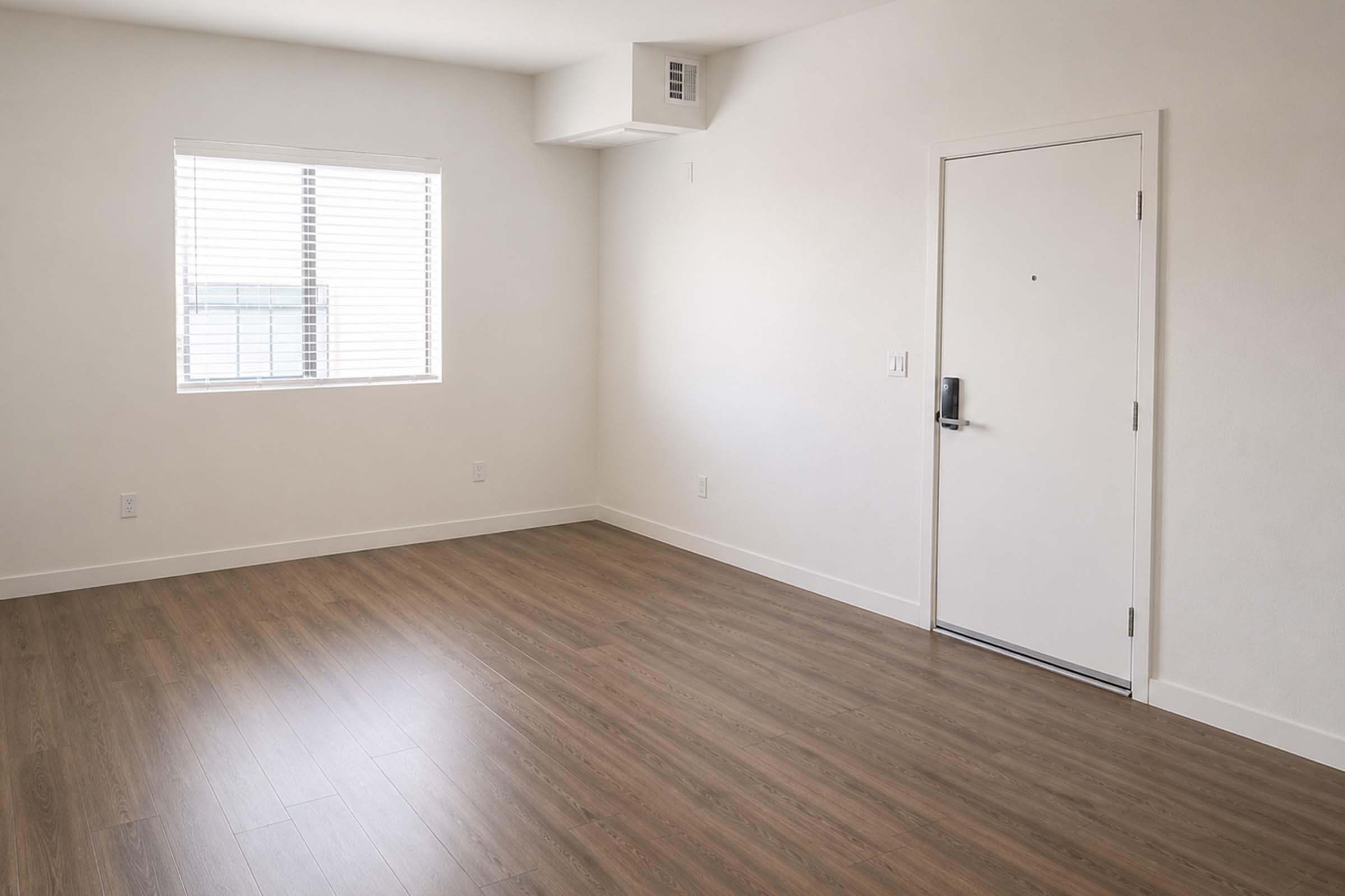 A spacious, empty room with light-colored walls and a wooden floor. There's a window with blinds on the left side and a white door on the right. The room is well-lit by natural light, creating a bright and airy atmosphere, suitable for various furnishings and designs.