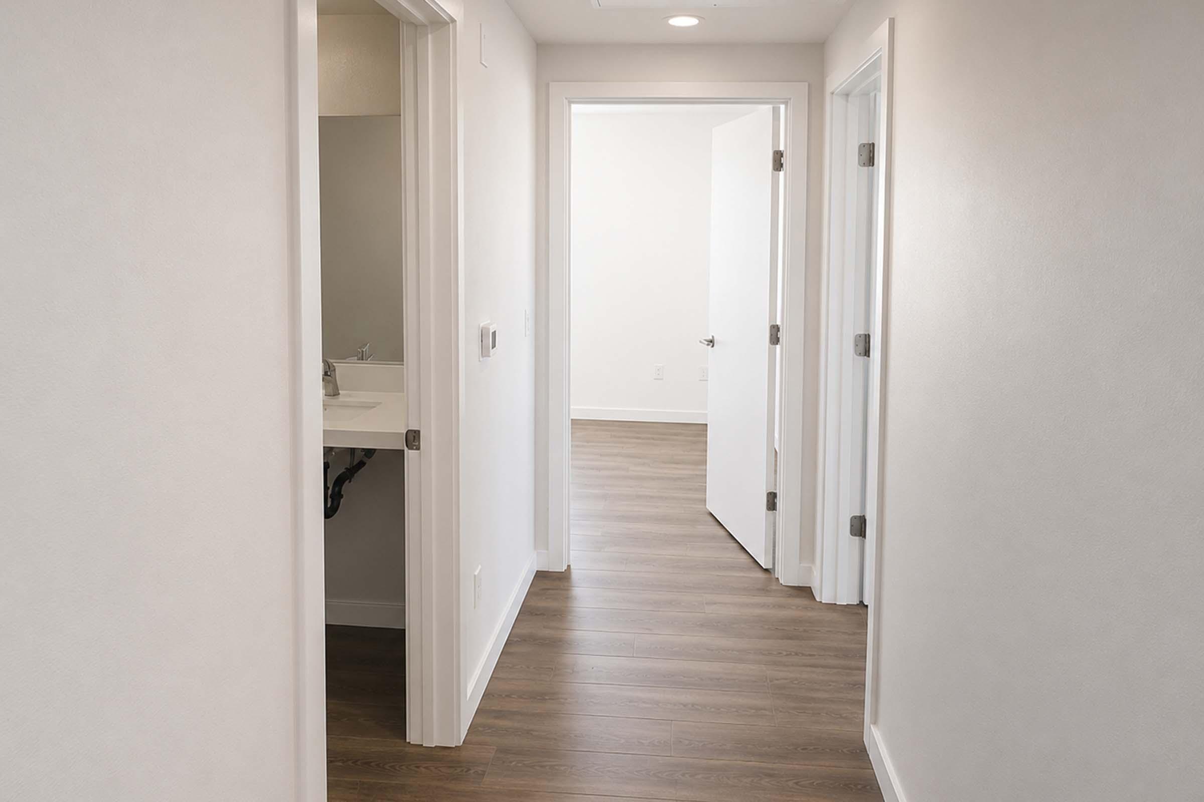 A narrow hallway with light-colored walls and wooden flooring. Two doors on the left lead to rooms, while a third door at the end of the hallway opens into another space. Natural light illuminates the area, creating a bright and minimalist atmosphere.