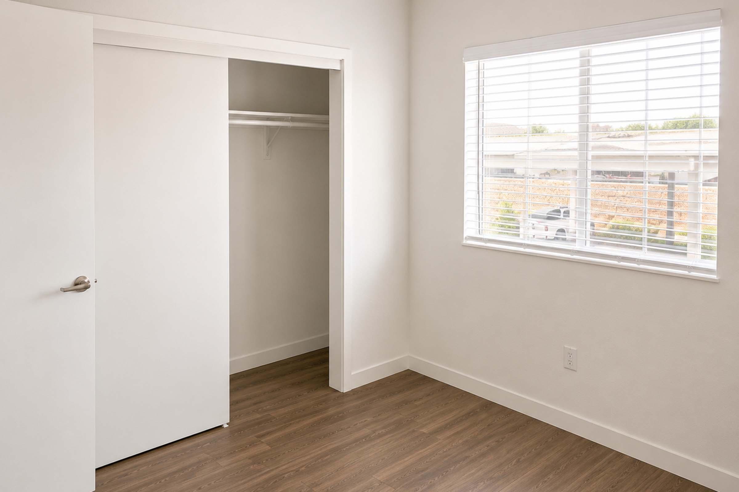 A well-lit, empty room featuring light-colored walls, a window with blinds, and a closet with sliding doors. The floor is wood-like, and natural light is coming in from the window, creating a spacious feel.