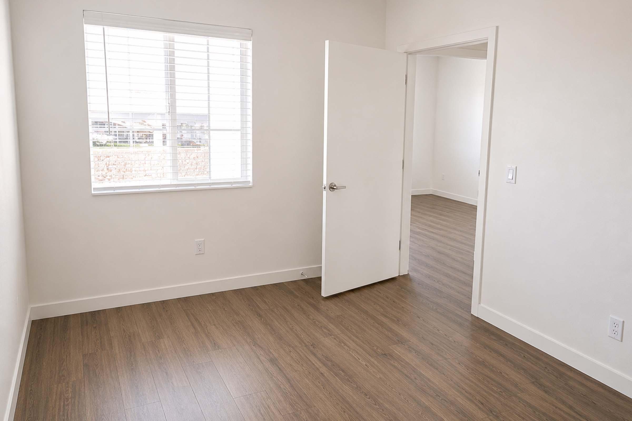 A bright, empty room with light-colored walls and a wooden floor. There is a window with blinds letting in natural light, a door slightly ajar leading to another room, and minimal furnishings, creating a spacious and airy atmosphere.