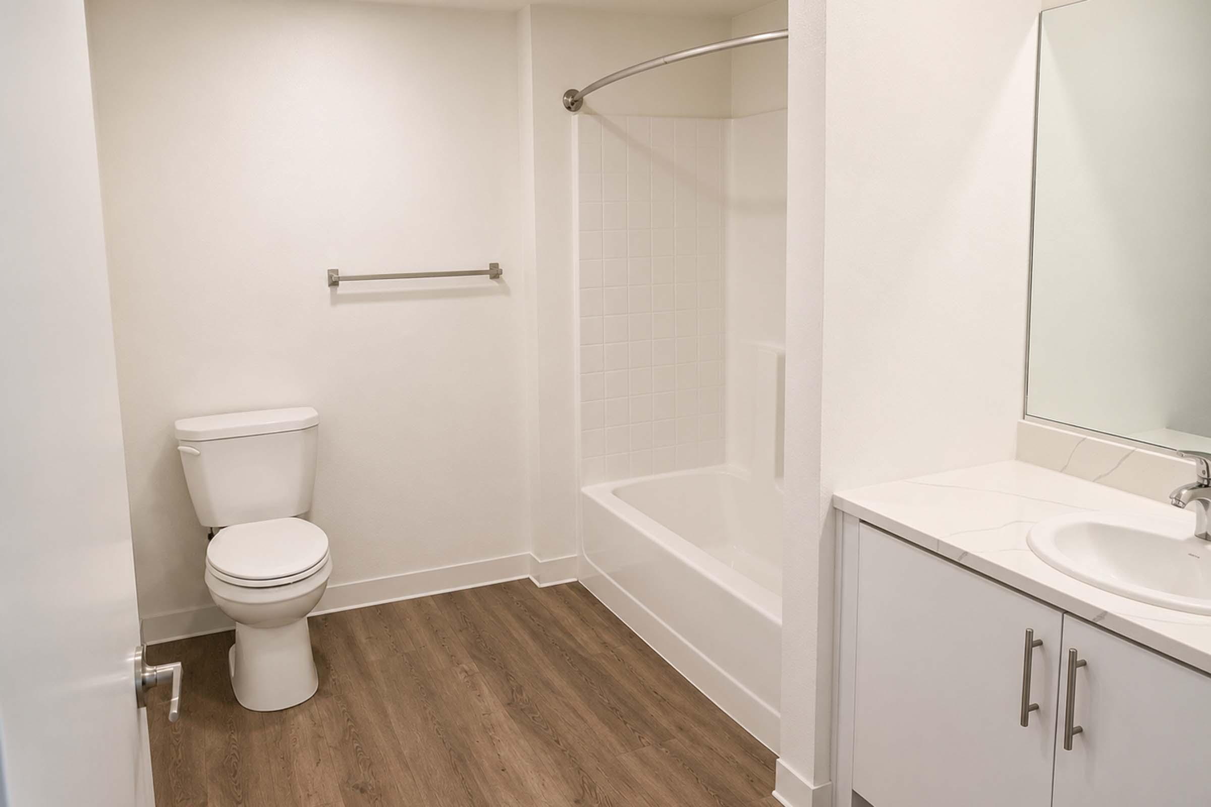 A clean, modern bathroom featuring a white toilet, a bathtub with a shower curtain, and a sink with a mirror above it. The walls are painted a light color, and the floor is made of wood-like tiles. A towel bar is mounted on the wall beside the bathtub.