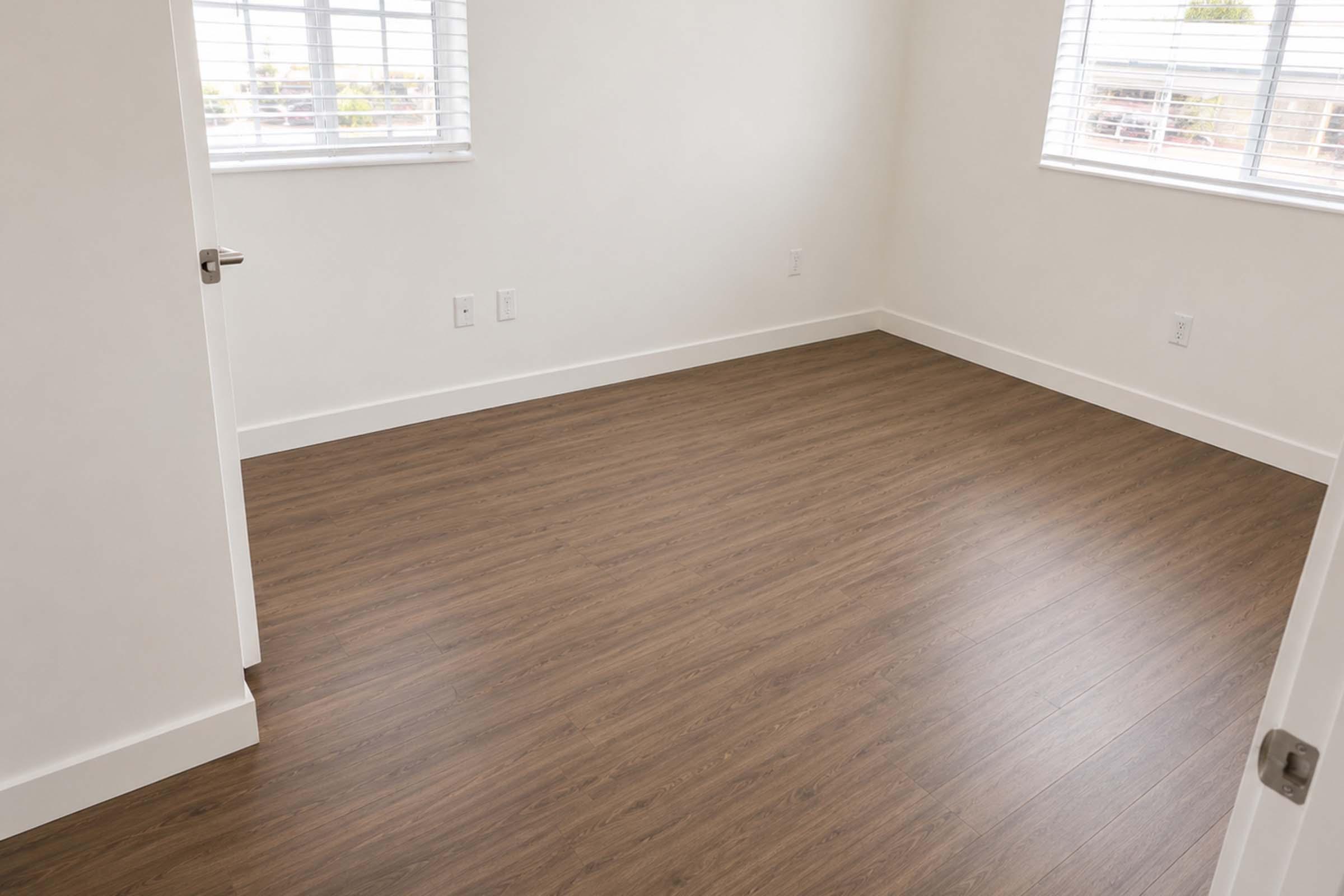 A vacant room with light-colored walls and wooden flooring. There are two windows with white blinds letting in natural light. A closed door is visible on the left side, and the room appears clean and unfurnished.