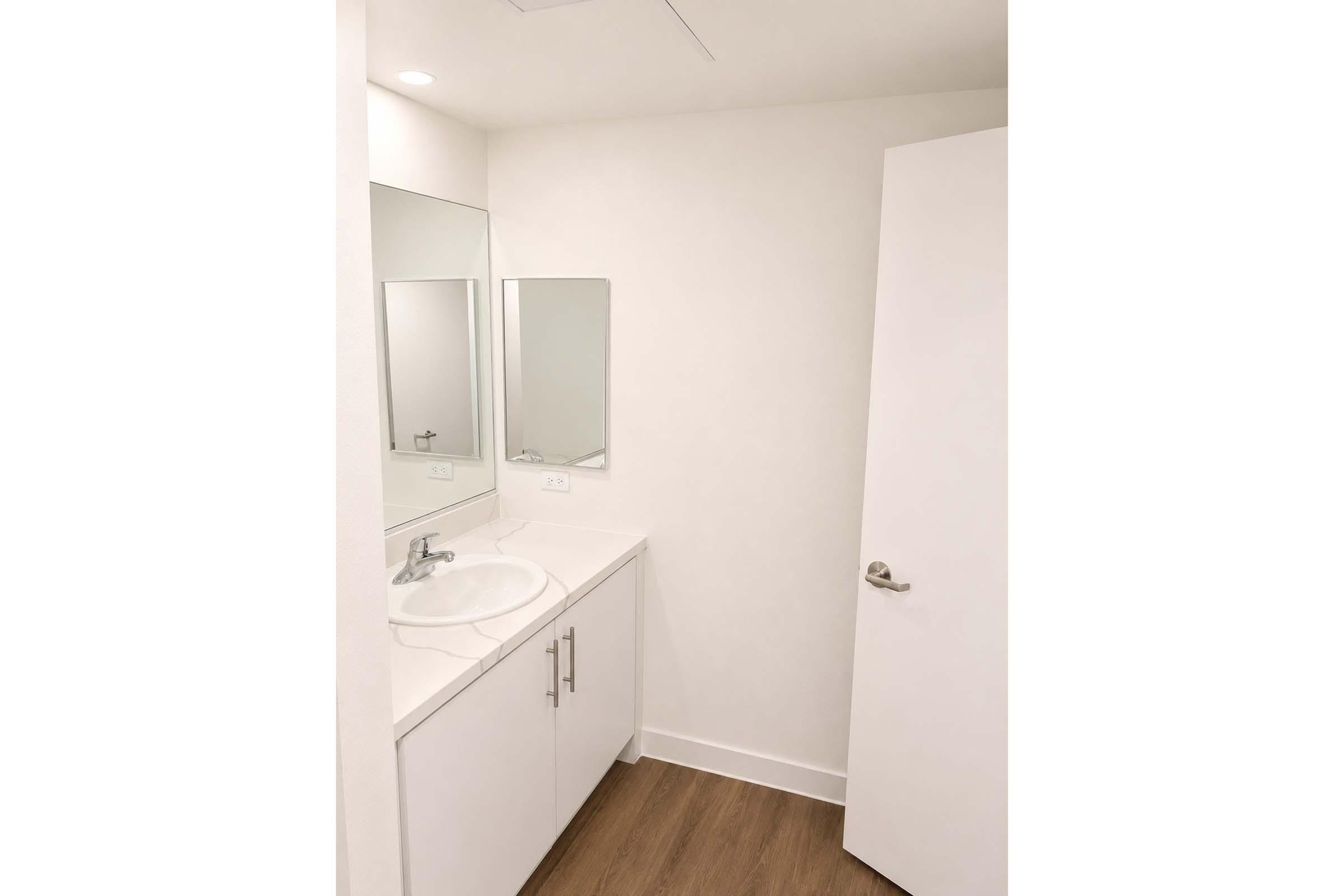 A modern bathroom featuring a white countertop with a sink, two mirrors above the sink, and a clean white cabinet below. The walls are painted in a light color, and there is a white door leading out of the room. The floor is made of wooden planks, adding warmth to the bright space.