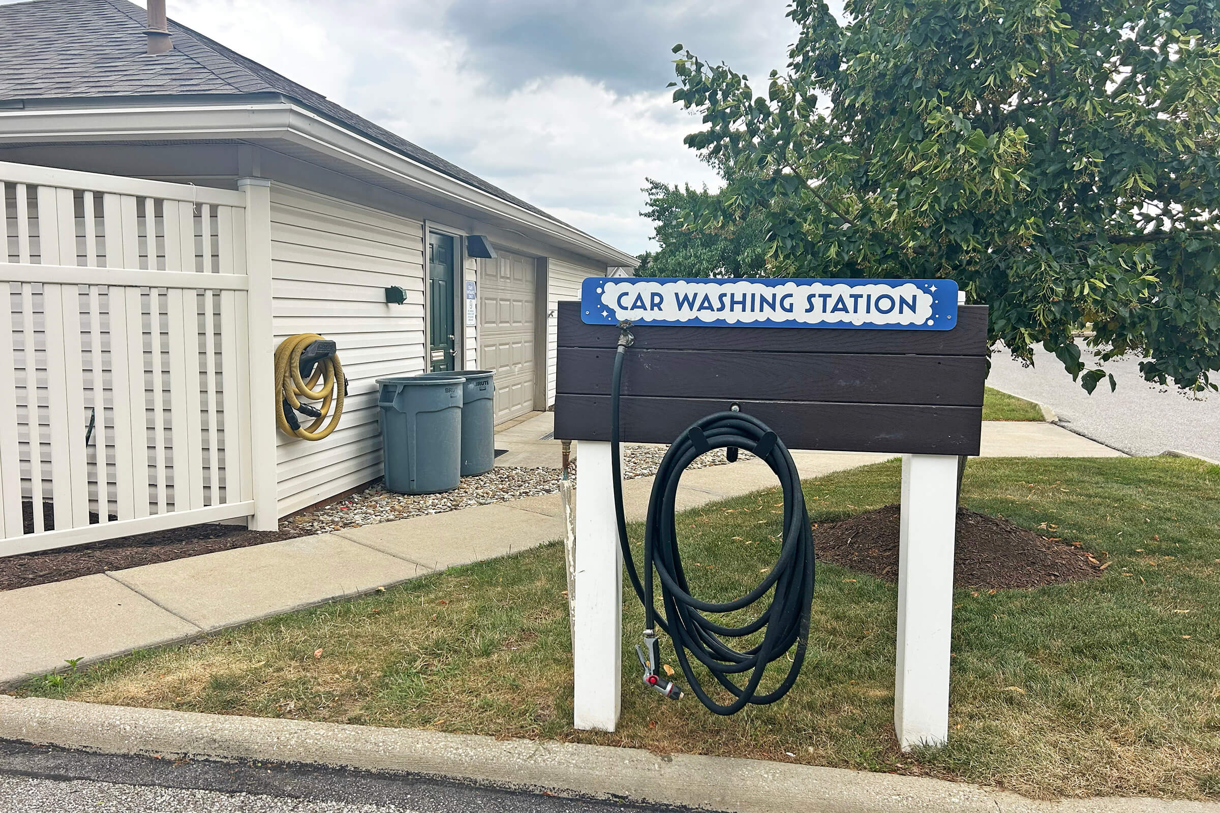Sign indicating a car washing station, with a mounted hose and nozzle hanging nearby. The area features a grassy patch and a light-colored building in the background, along with a trash can. The sky is partly cloudy.