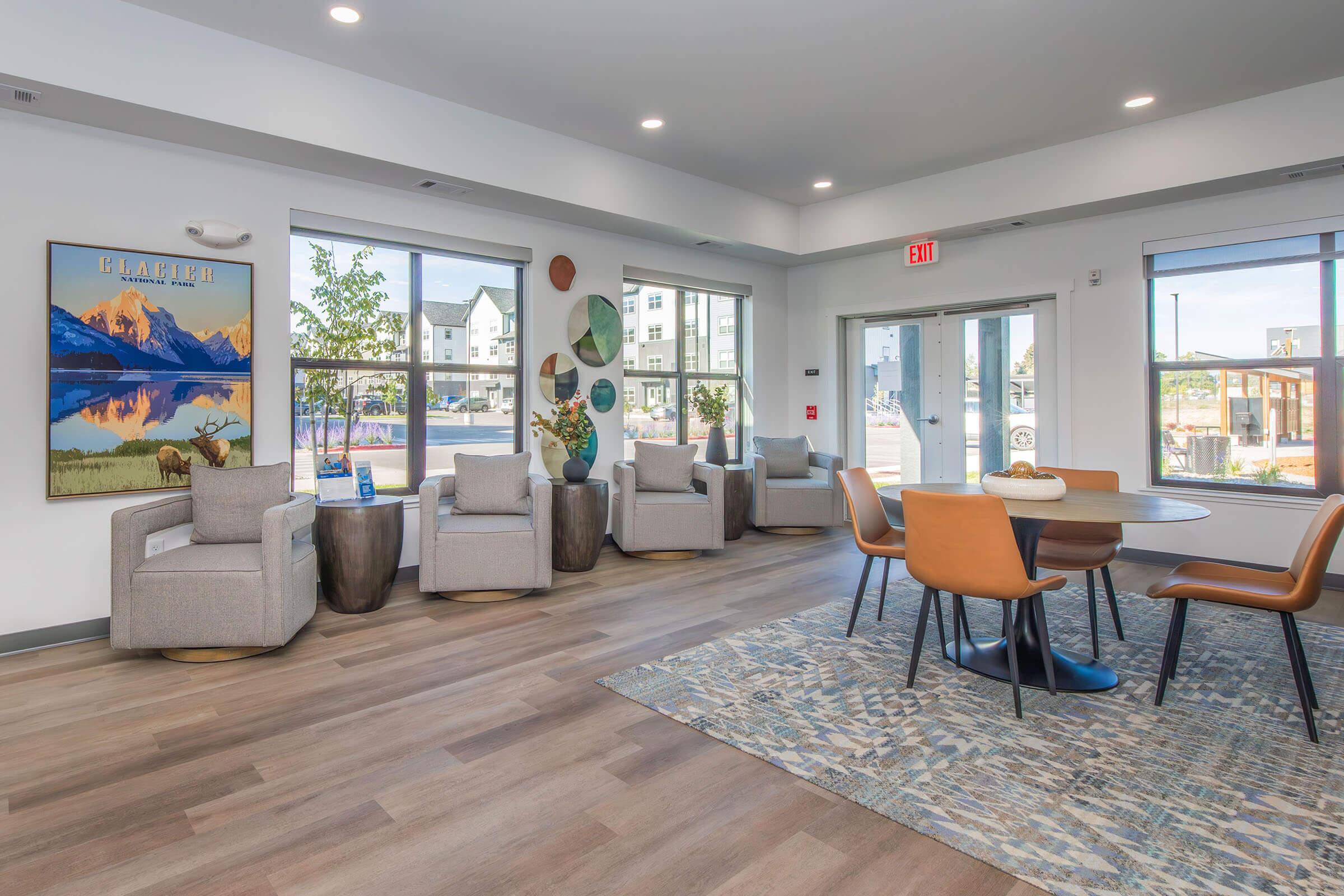 A modern lounge area featuring light gray armchairs and wooden side tables. A round dining table with orange chairs is in the center. Large windows provide natural light and outdoor views. There is a decorative wall art of mountains and trees, enhancing the welcoming atmosphere.