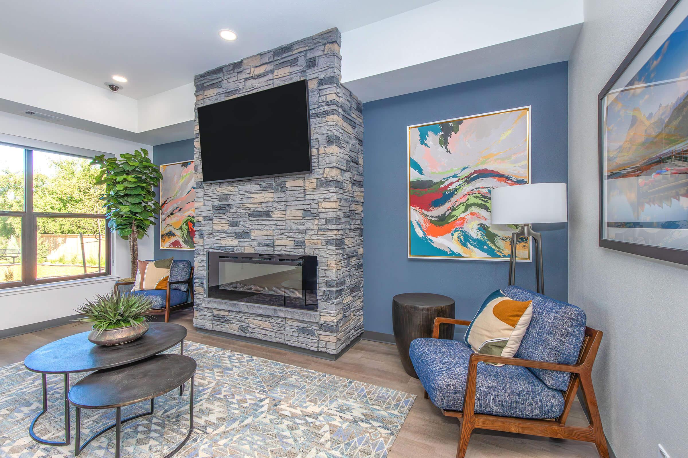 A modern living room featuring a stone fireplace with a mounted flat-screen TV. Two comfortable blue armchairs with colorful cushions are positioned around a round coffee table. A vibrant area rug adds texture, and there is a tall green plant in the corner. Large windows provide natural light.