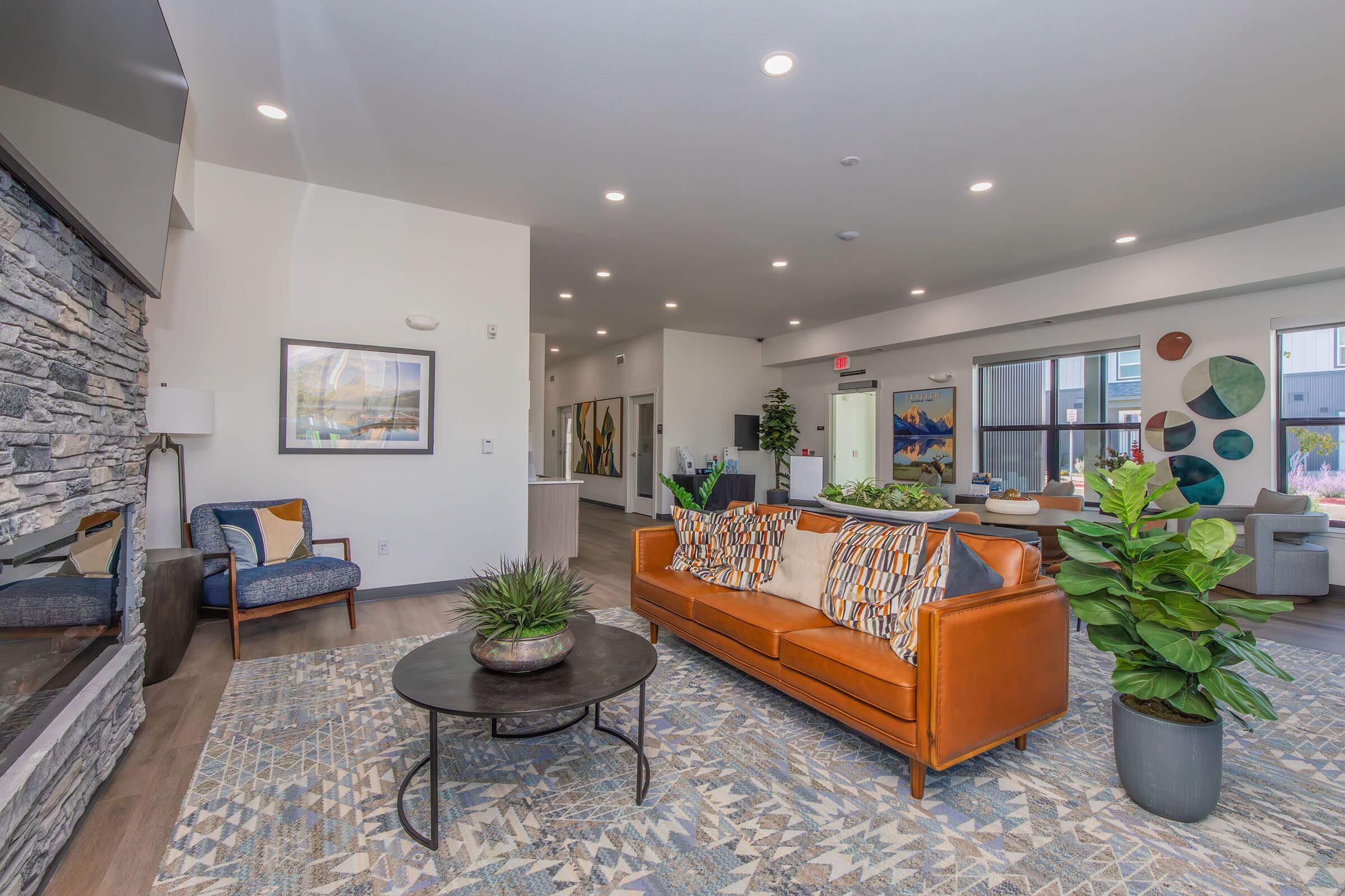 A modern living room with a cozy brown leather sofa adorned with colorful cushions, a round coffee table with a plant, and a stone fireplace. There are decorative chairs and art pieces on the walls, with bright lighting and an open layout leading to other areas, creating a warm and inviting atmosphere.