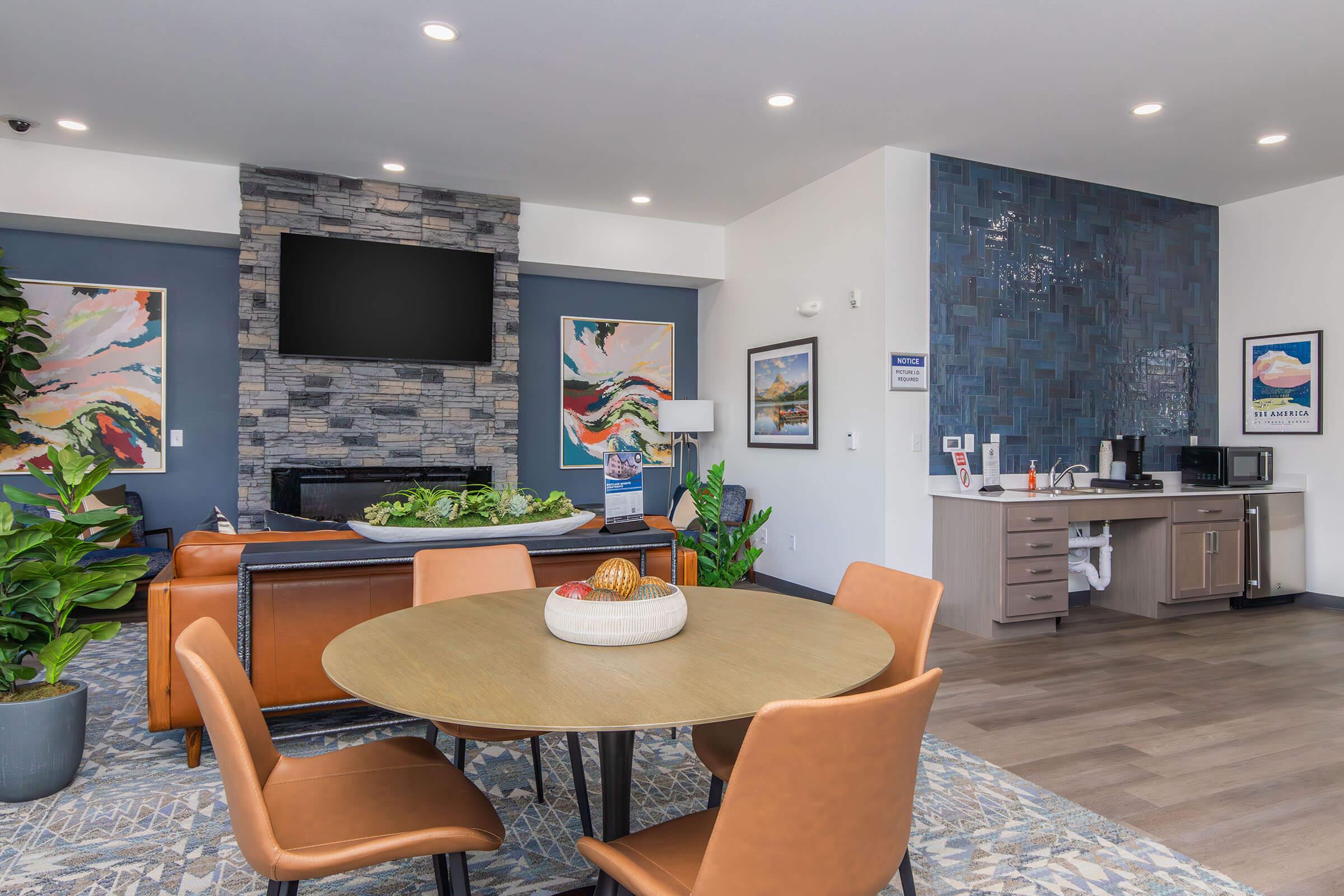 A modern lounge area featuring a round table with orange chairs, a stone fireplace, and a large wall-mounted TV. The decor includes colorful abstract art, indoor plants, and a kitchenette with a microwave, creating a welcoming and stylish environment.