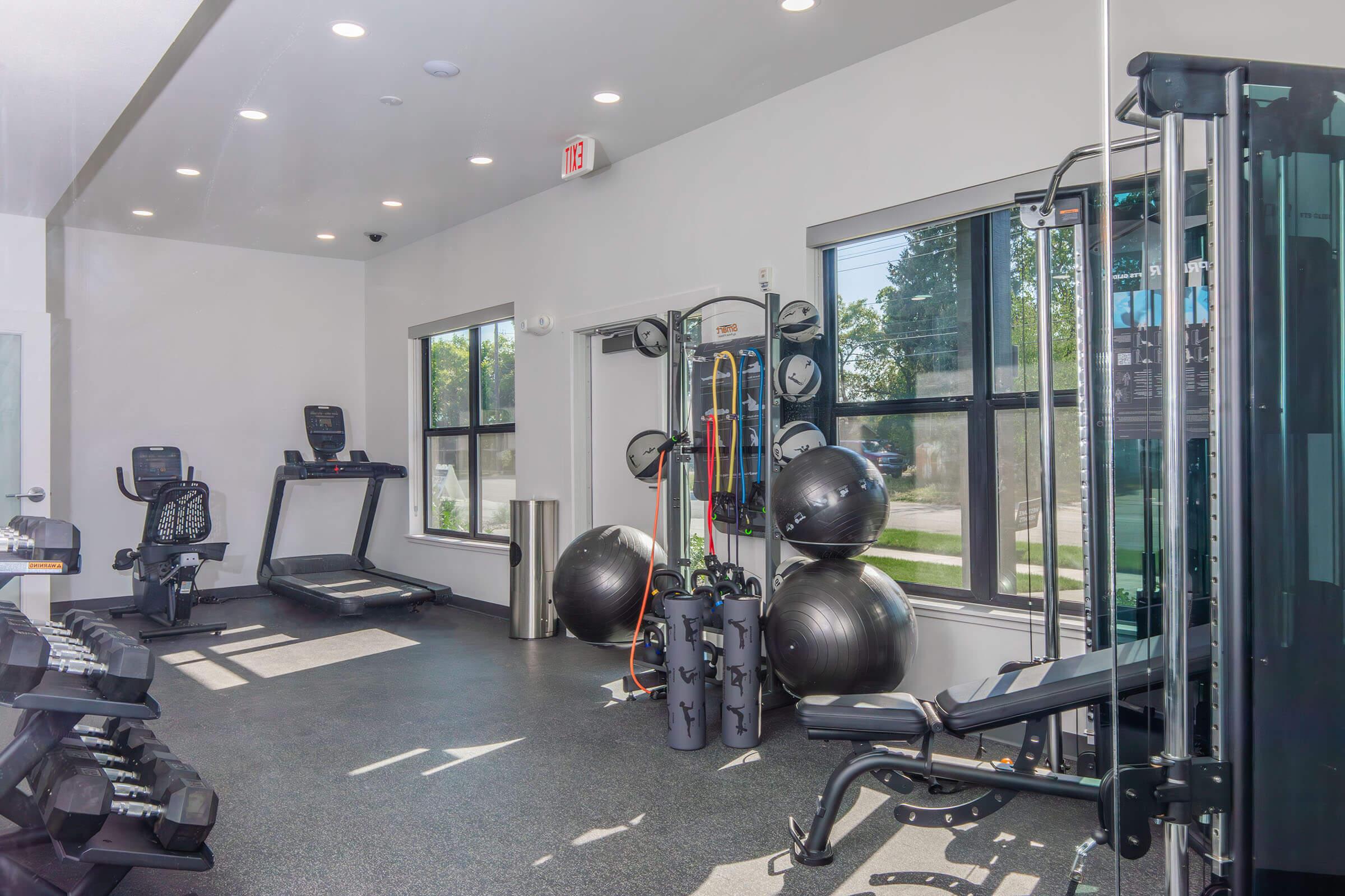 A bright and modern gym interior featuring various exercise equipment including a treadmill, weight bench, dumbbells, and resistance bands. Large windows allow natural light, enhancing the open feel. The space is clean and well-organized, ideal for workouts.