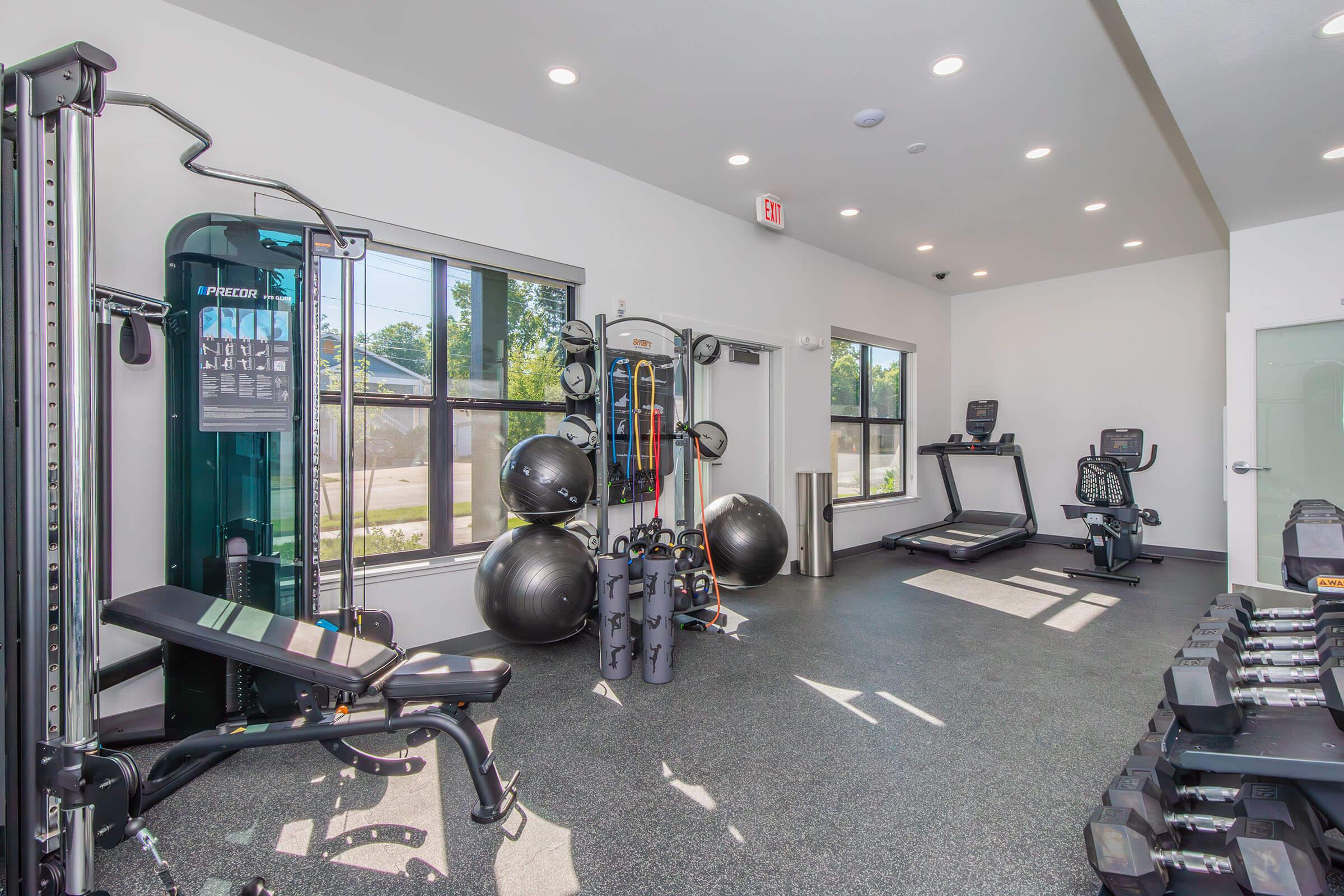 A modern gym interior featuring various workout equipment, including a multi-station exercise machine, free weights, a treadmill, and stability balls. The space has large windows allowing natural light in and a sleek, clean design with rubber flooring.