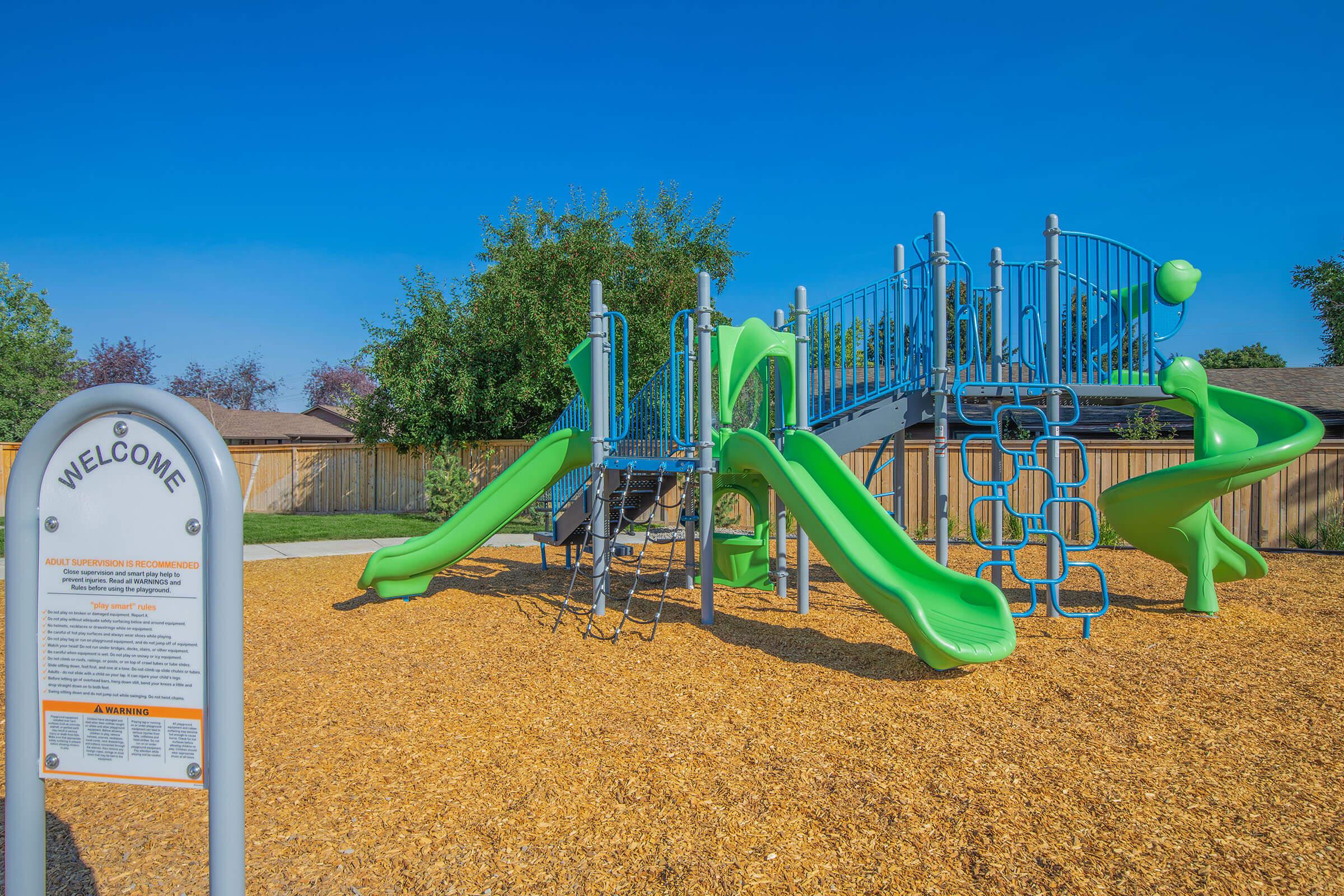 Playground equipment with green and blue slides, climbing structures, and monkey bars surrounded by a wood chip surface. In the foreground, a sign welcomes visitors to the area, with visible safety guidelines and information. The scene is set under a clear blue sky with trees in the background.