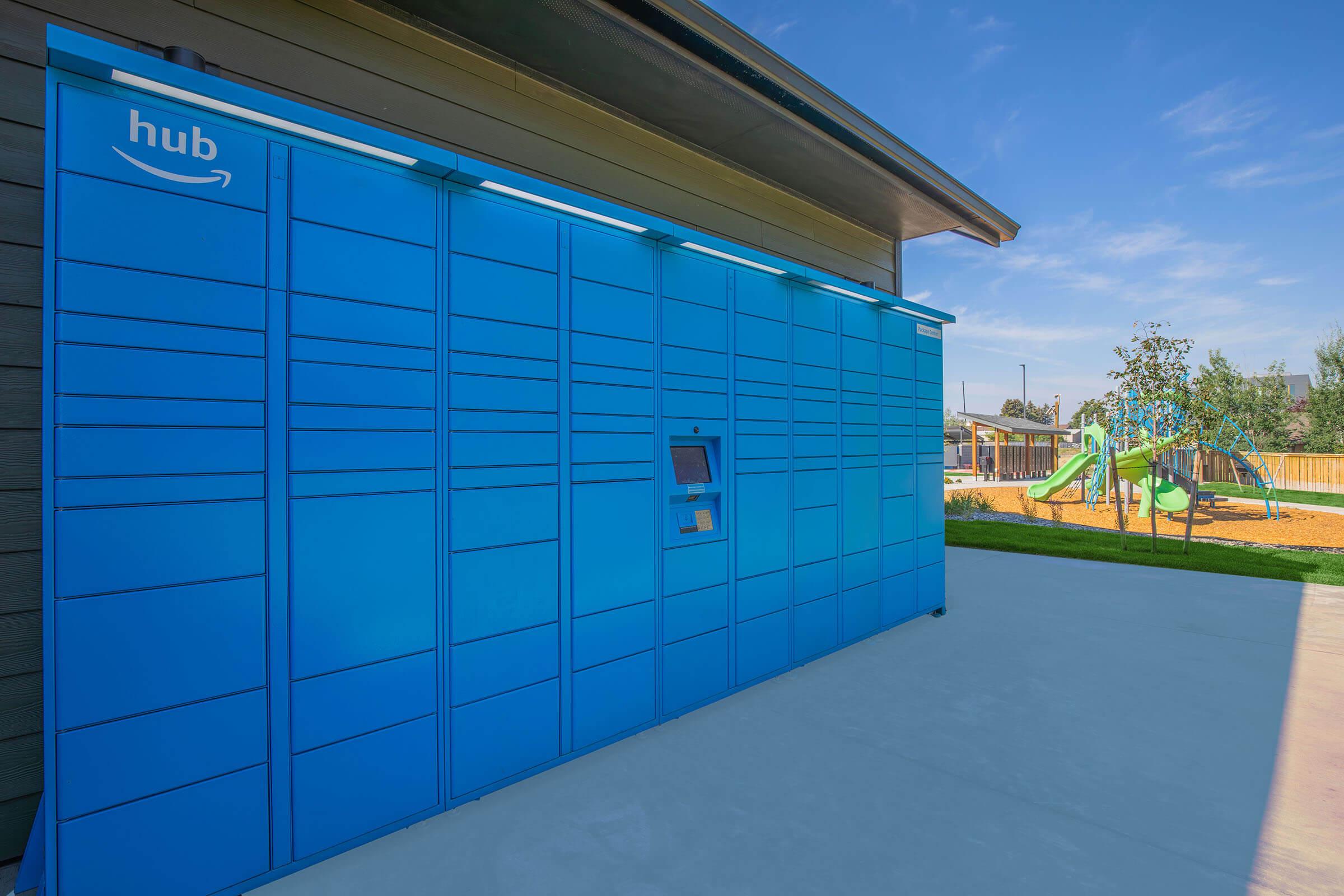 A blue Amazon Hub locker is placed against a building, providing secure package delivery. In the background, there is a playground with green equipment and a clear blue sky above. The scene is well-lit and showcases a modern, outdoor setting.