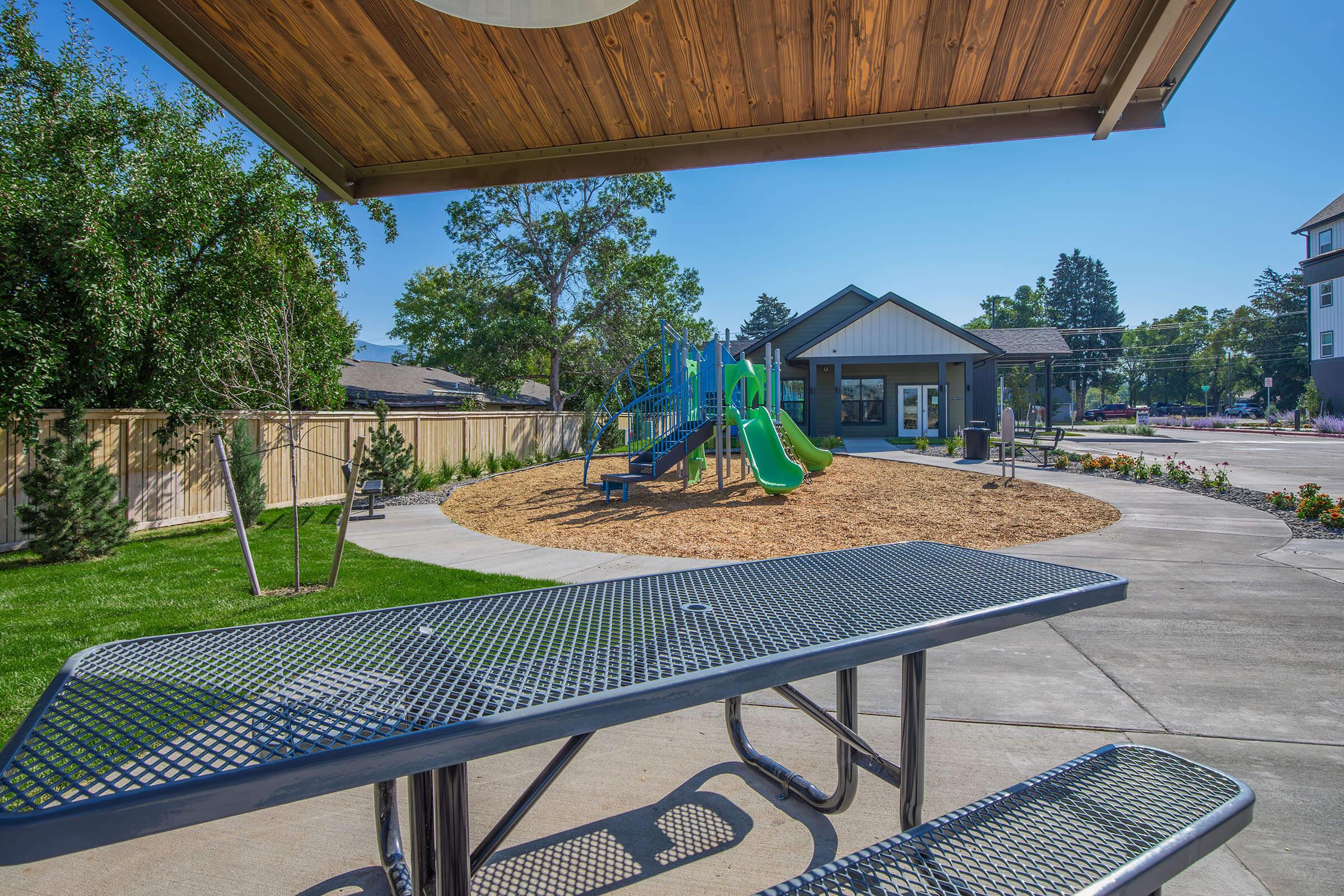 A view of a playground featuring green slides and climbing structures, surrounded by mulch and grass. In the foreground, a metal picnic table is visible under a wooden shaded area. The playground is part of a landscaped area with trees and a building in the background.