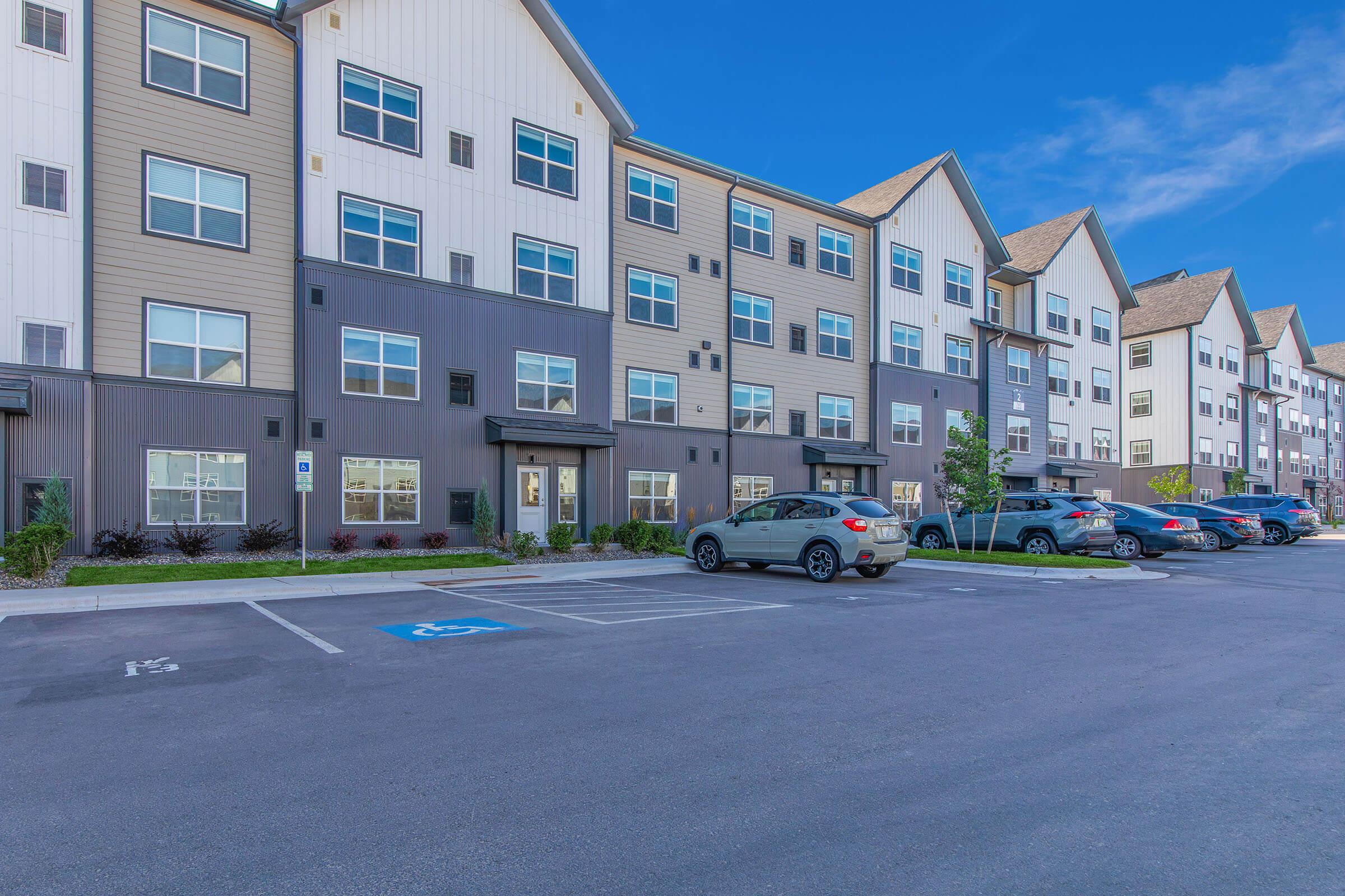A modern multi-story apartment building with a mix of exterior finishes, featuring large windows and a well-maintained parking lot. Several parked cars are visible, along with designated handicapped parking spaces and green landscaping in front of the building. The sky is clear and blue.