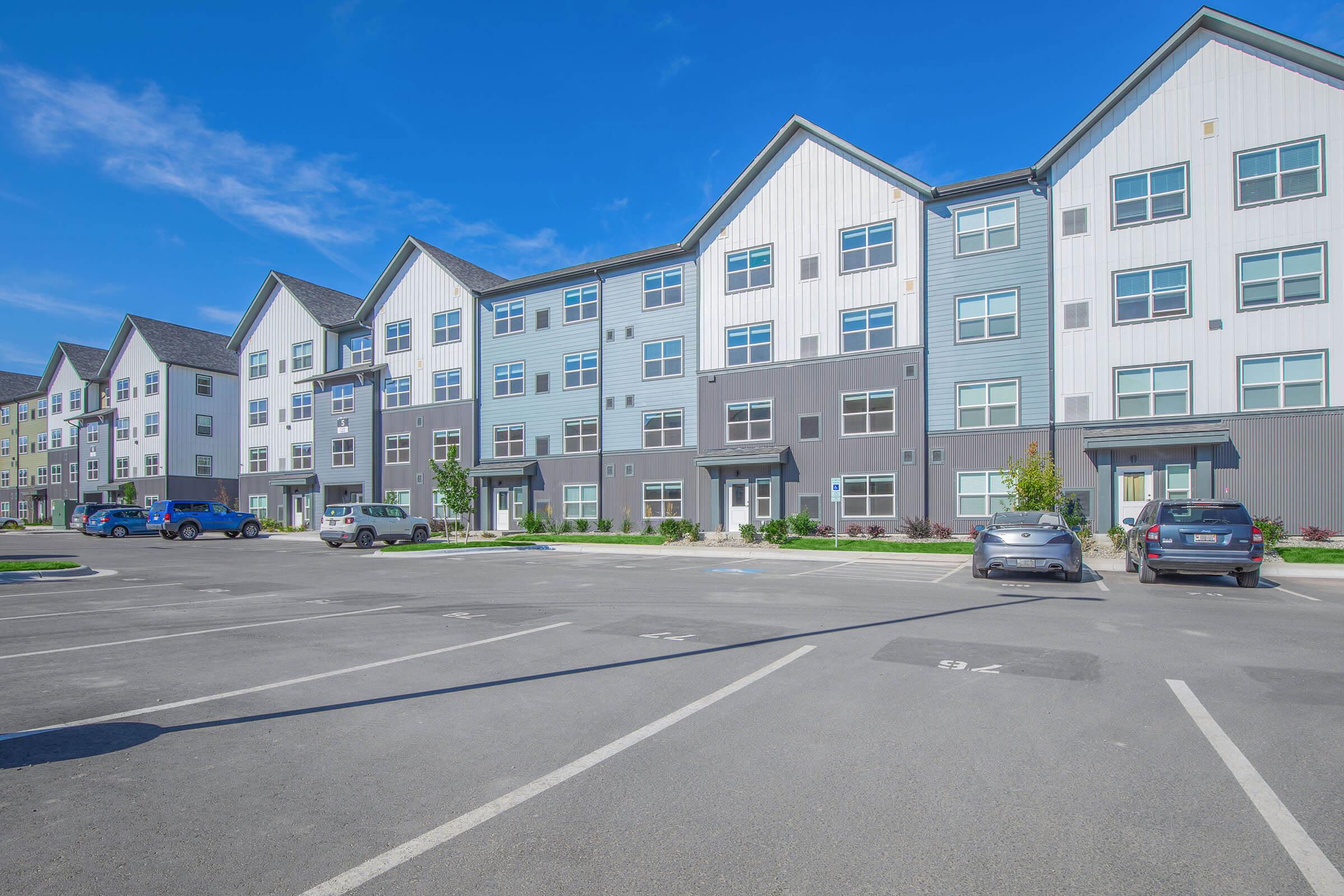 Modern multi-story apartment building with a mix of light and dark siding. The building features large windows and multiple entrances. A parking lot in front has several parked vehicles, and there are landscaped areas with greenery. The sky is clear and blue, indicating a sunny day.