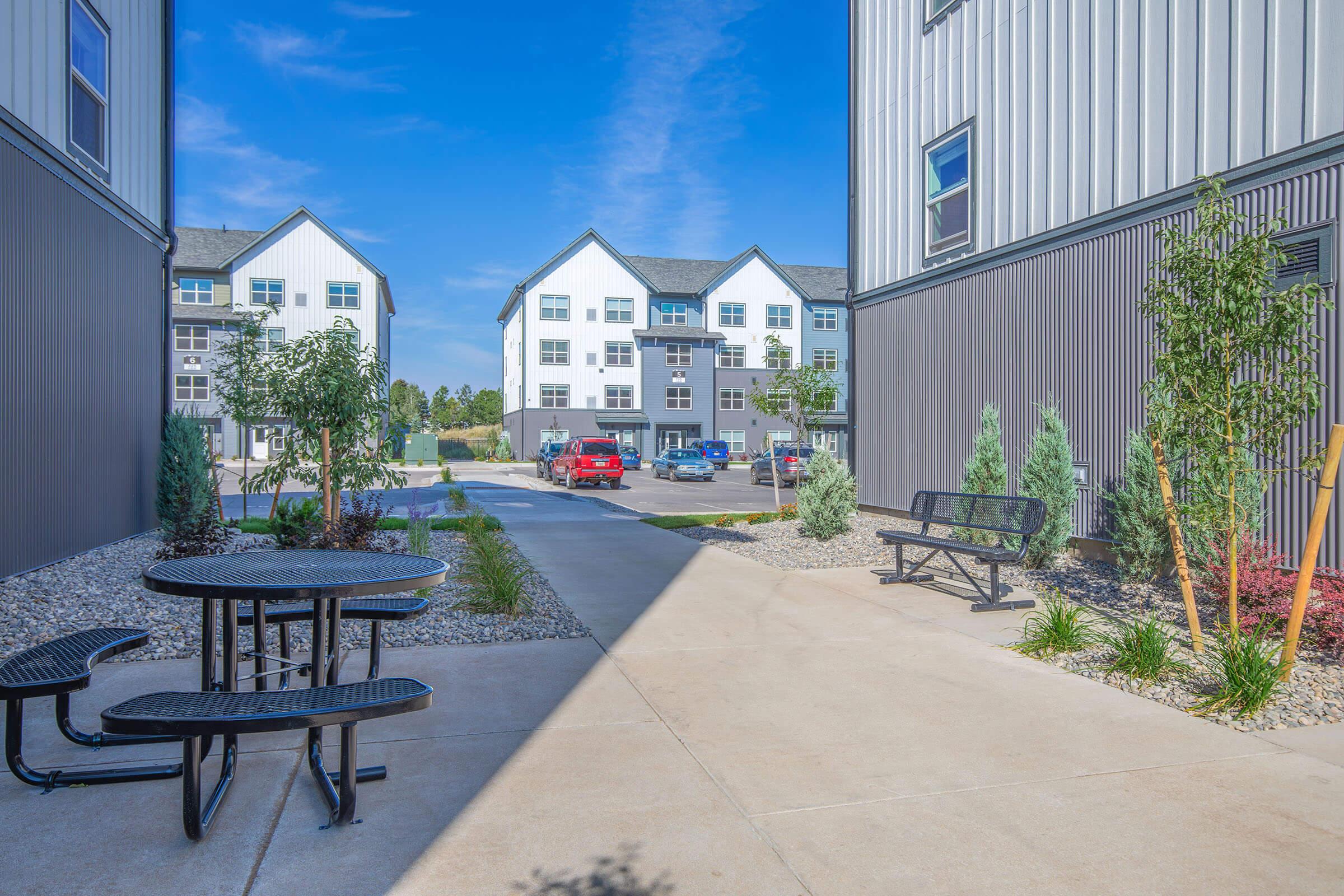 A modern residential complex featuring a landscaped courtyard with picnic tables, greenery, and a pathway. The view shows multi-story buildings in a contemporary style, with cars parked nearby and a clear blue sky overhead.