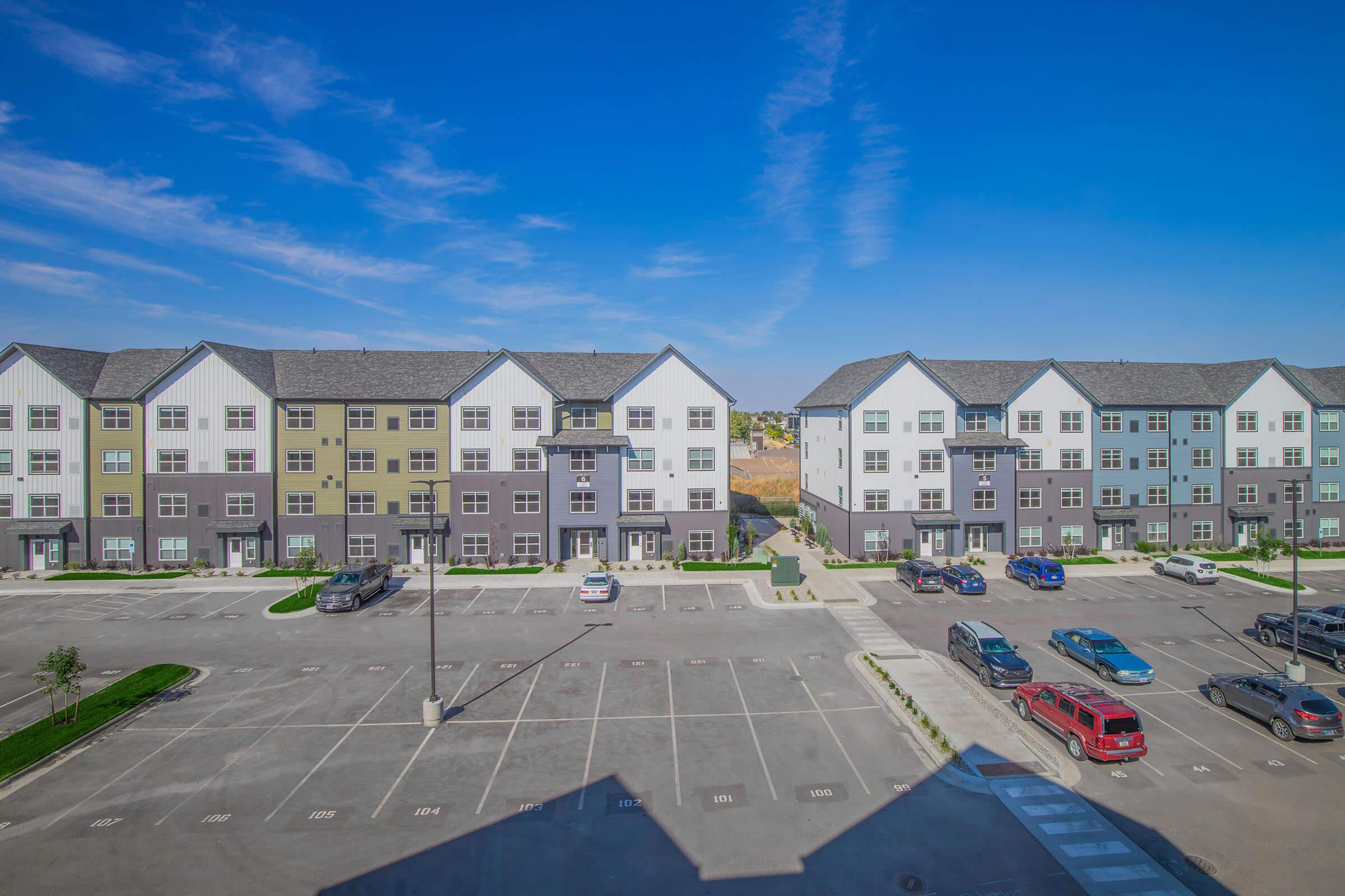 A view of modern multi-story apartment buildings with a mix of green, gray, and white exteriors, surrounded by a parking lot. Several cars are parked in the lot, and the sky is clear with a few clouds, indicating a sunny day. The setting has landscaped areas with grass.
