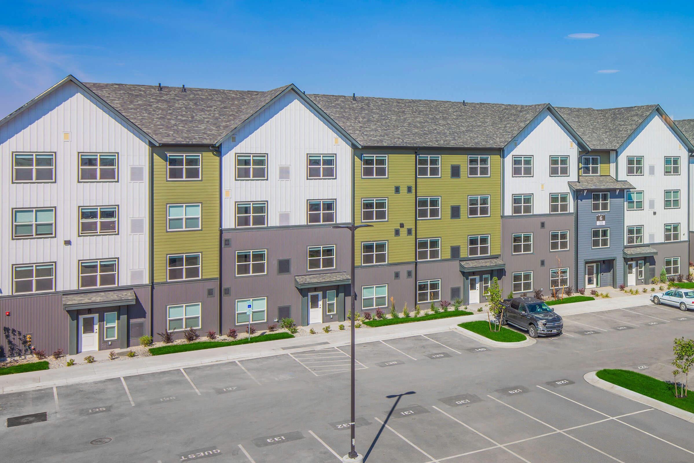 Modern multi-story apartment building with a mix of gray, green, and white facades. The structure features large windows and a contemporary design. In the foreground, there is a parking lot with several parked cars and landscaped areas with small plants. The sky is clear and blue.