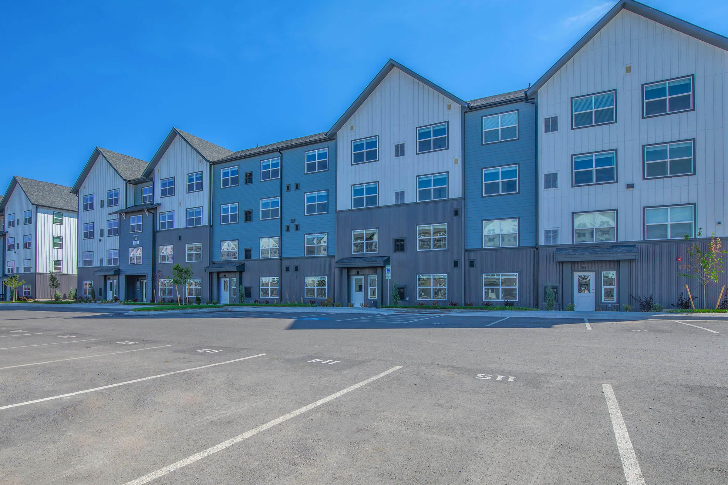 A modern, multi-story residential building with a mix of gray and white exterior. The structure features large windows and a gabled roof. In the foreground, there is an empty parking lot with marked spaces. Clear blue skies provide a bright backdrop.