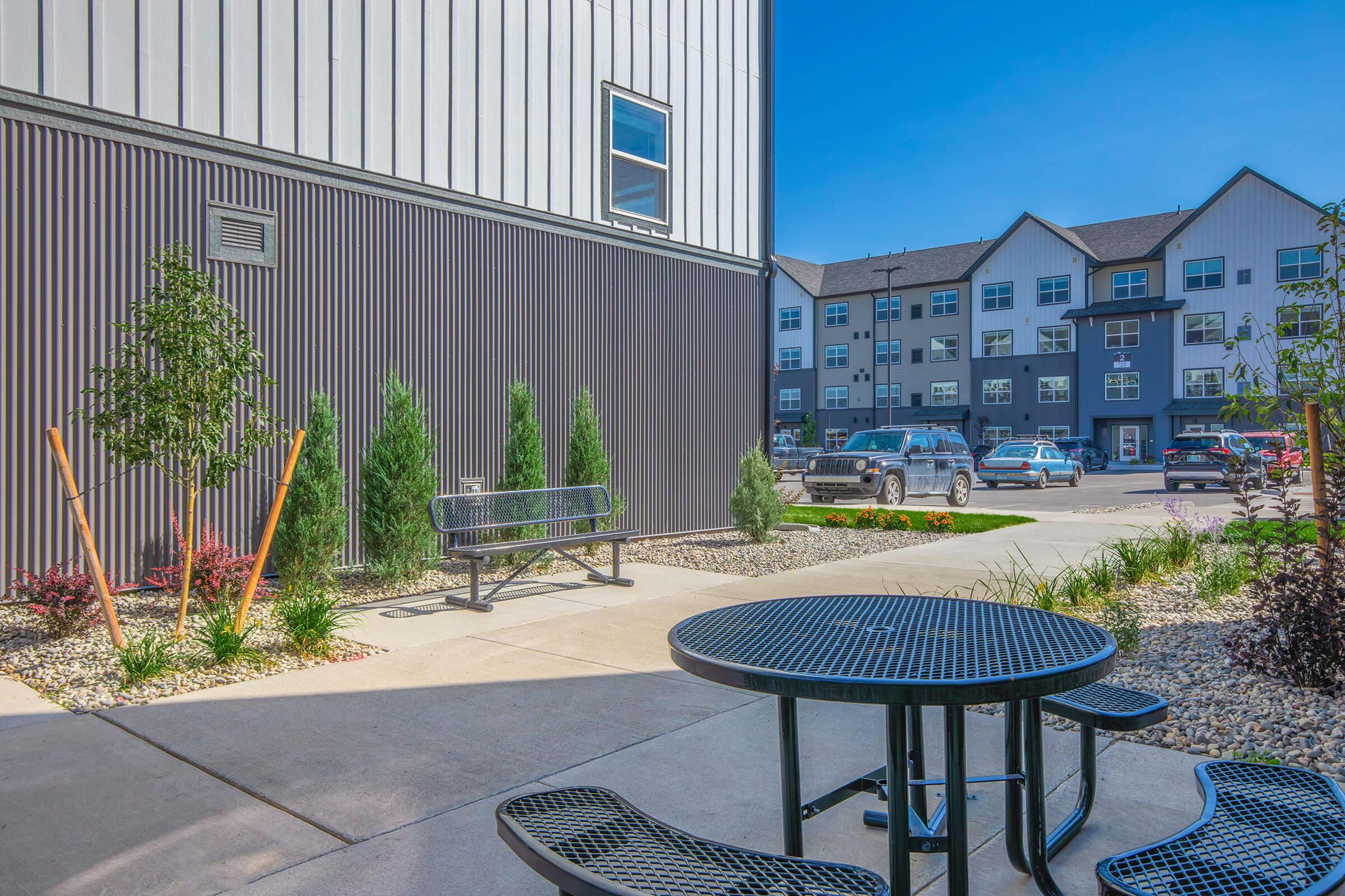 A modern outdoor courtyard featuring a round black picnic table with attached benches, surrounded by landscaped plants and small trees. In the background, contemporary apartment buildings are visible under a clear blue sky. There are additional benches and parked cars nearby.
