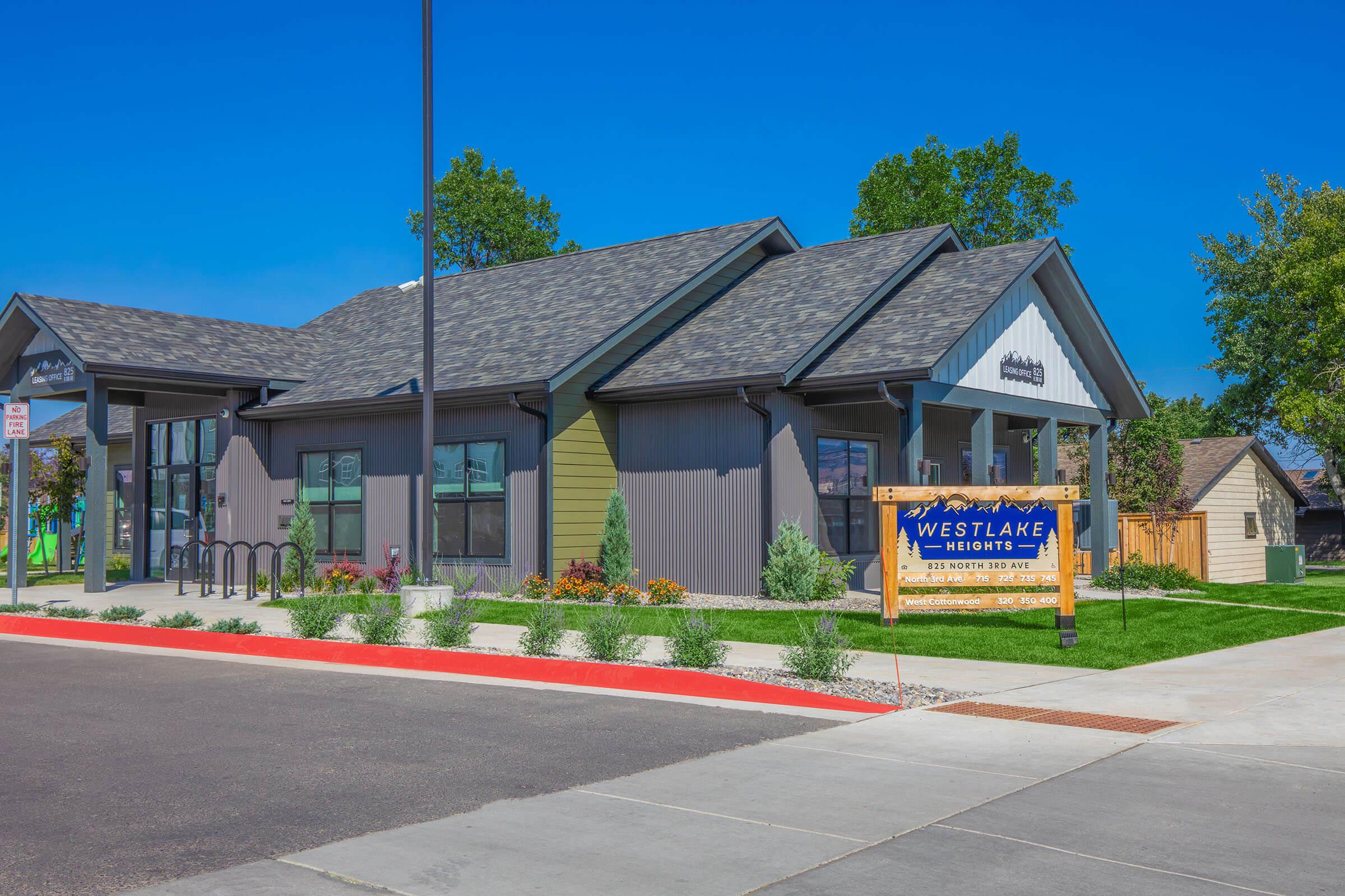 A modern building with a sloped roof and a combination of gray and green siding. It features large windows and a welcoming entrance. A prominent sign reading "Westlake" is displayed in front, along with well-maintained landscaping and a clear blue sky above.