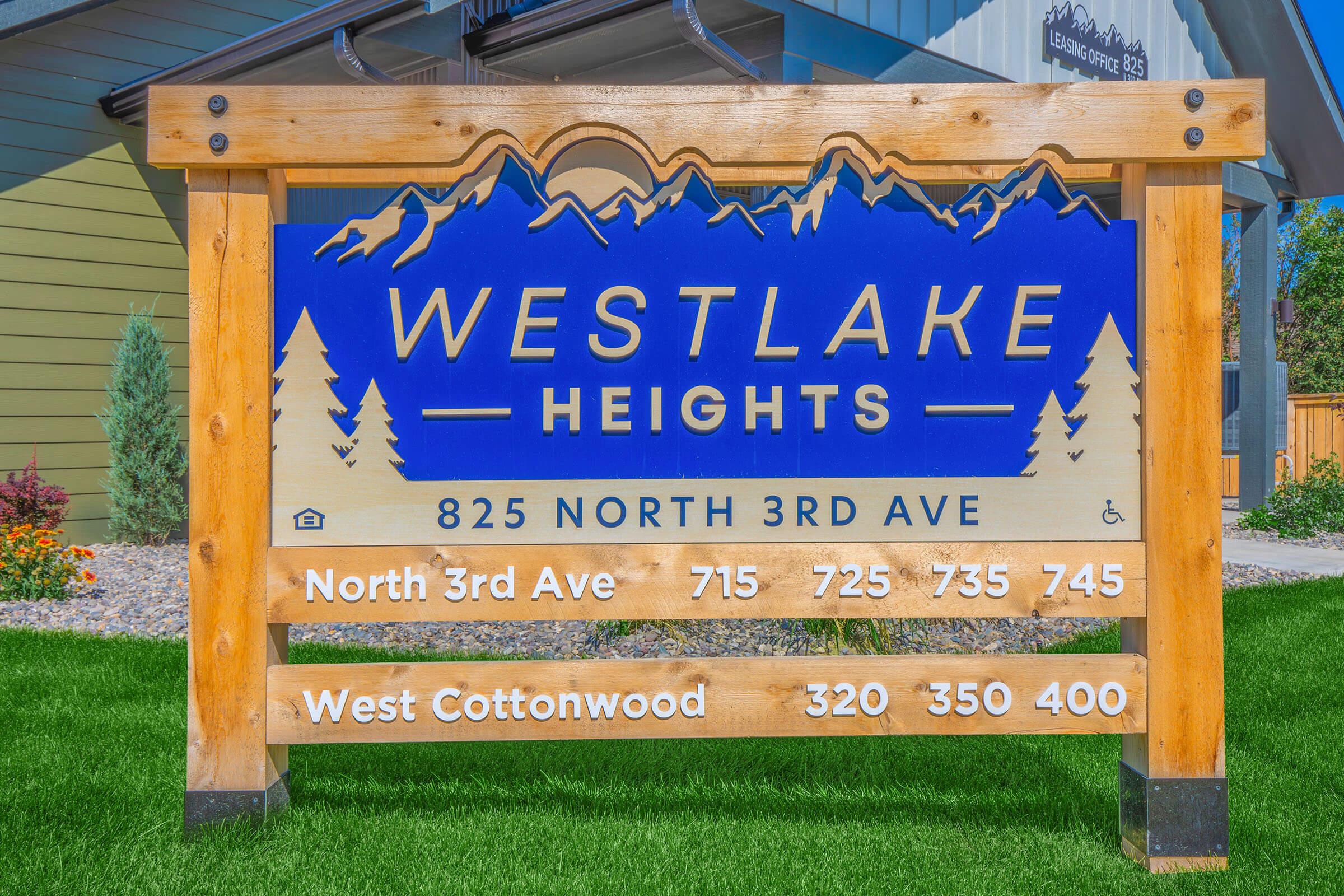 Sign for Westlake Heights featuring a blue background with white text. The sign includes the address "825 North 3rd Ave" and directional information with numbers for North 3rd Ave (715, 725, 735, 745) and West Cottonwood (320, 350, 400). Surrounding grass and plants enhance the setting.