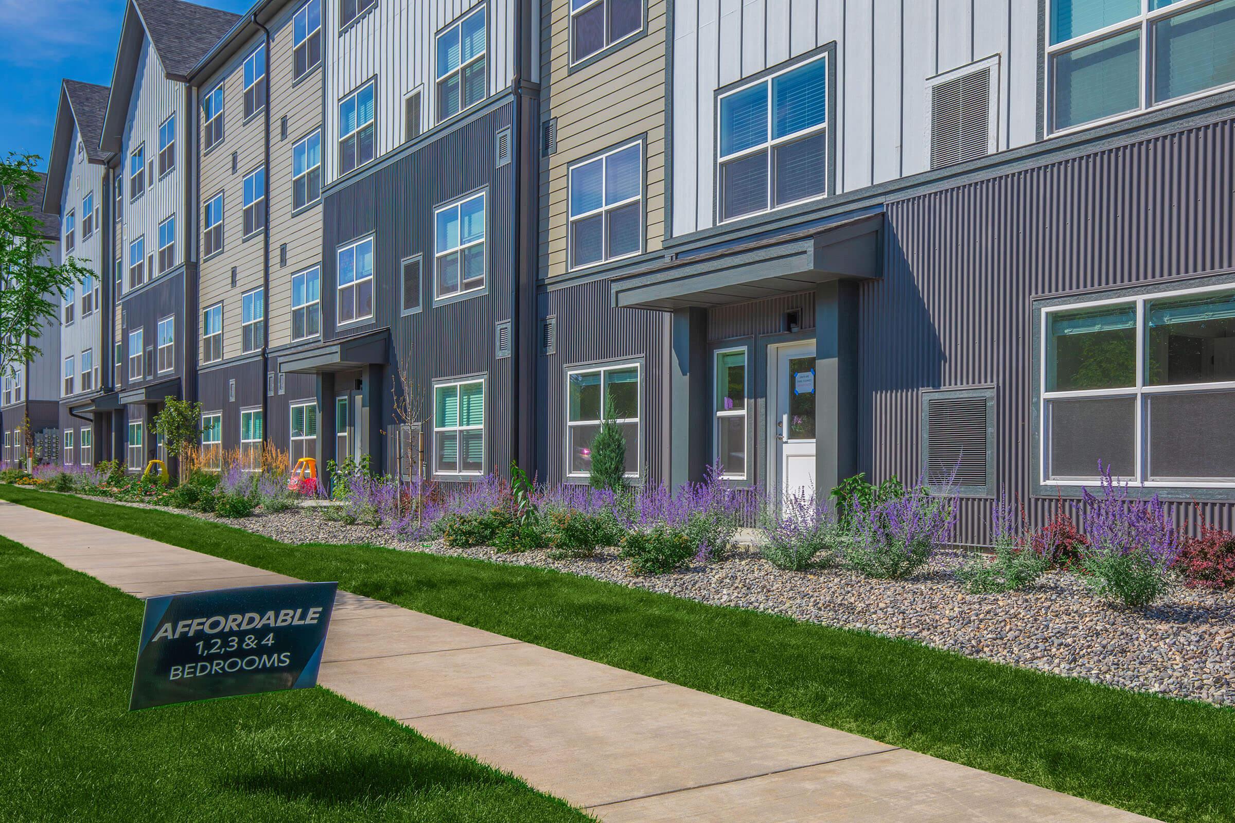 A well-maintained apartment complex with a modern exterior featuring a mix of siding colors. The building is set against a clear blue sky, with a landscaped path adorned with colorful flowers and greenery. A sign in front advertises affordable 1, 2, 3, and 4 bedroom options.