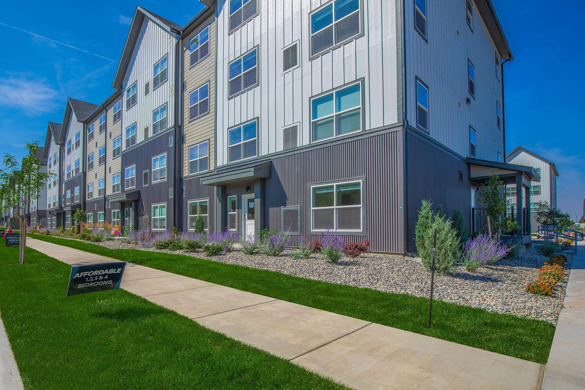 Modern multi-story apartment building with a sleek design. The exterior features a mix of metal and siding materials. A well-maintained pathway leads up to the entrance, surrounded by landscaped greenery and colorful flowers. A sign prominently displays “Affordable Bedrooms.” Clear blue sky in the background.