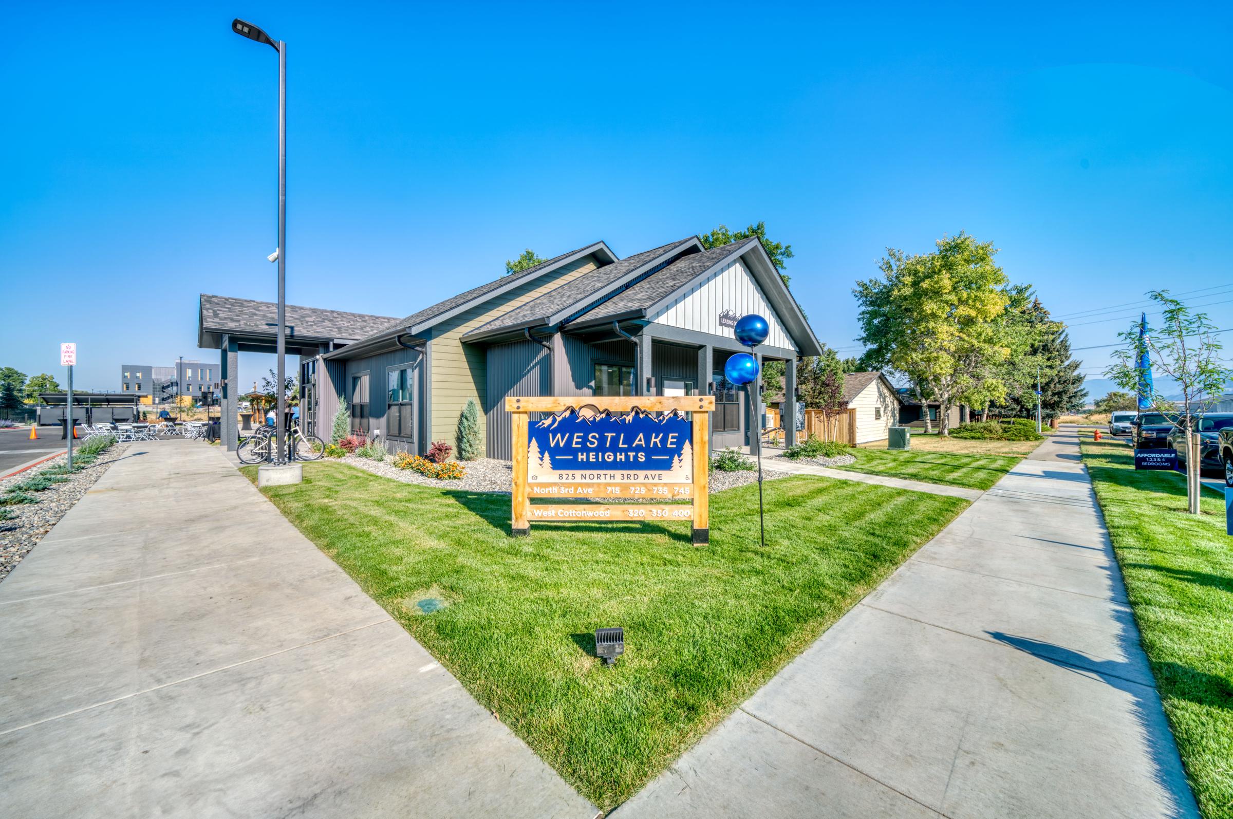 A modern building with a sign reading "Westlake Heights" in front, surrounded by well-manicured grass and decorative landscaping. The scene features clear blue skies and pathways leading to the entrance, with a few trees and light poles visible in the area.