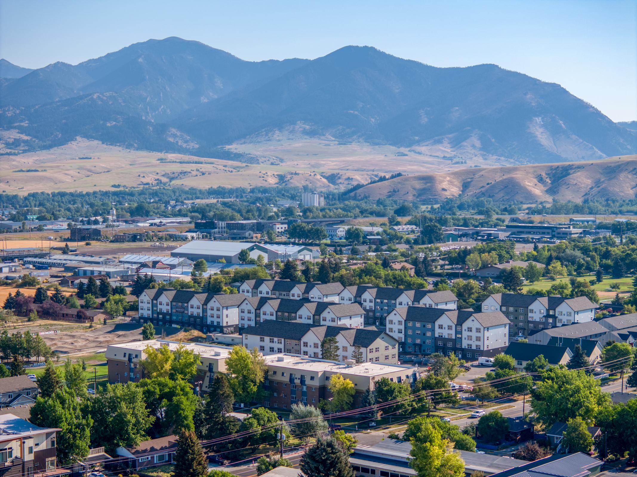A panoramic view of a residential area with several multi-story buildings, surrounded by trees and open fields, set against a backdrop of mountains under a clear blue sky. The landscape features a mix of urban and natural elements, highlighting the contrast between housing and the surrounding terrain.