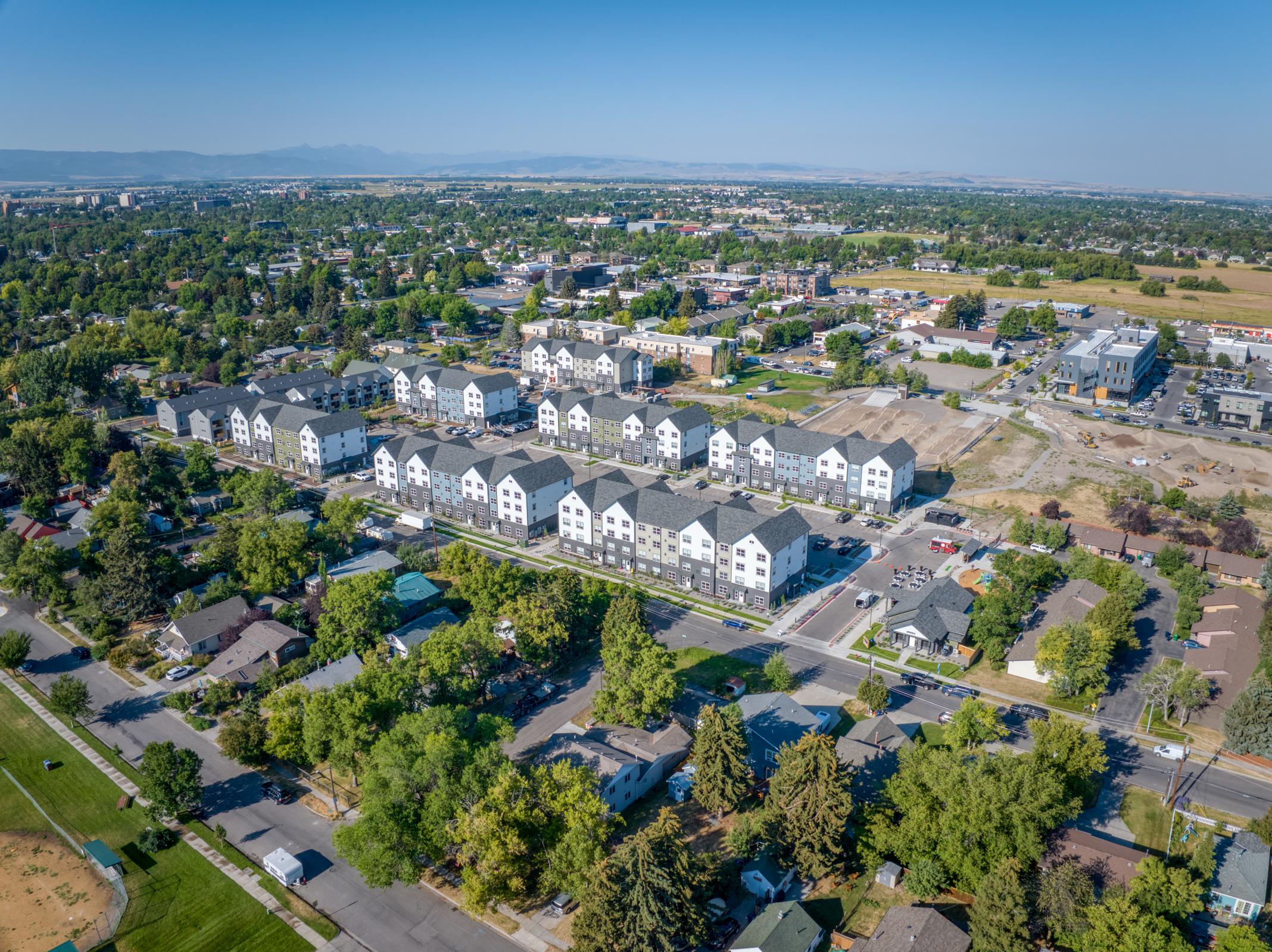 Aerial view of a suburban area featuring a row of modern residential buildings, green trees, and roads. In the background, there are commercial buildings and open land, with distant mountains visible on the horizon. The scene captures a mix of residential and urban development.