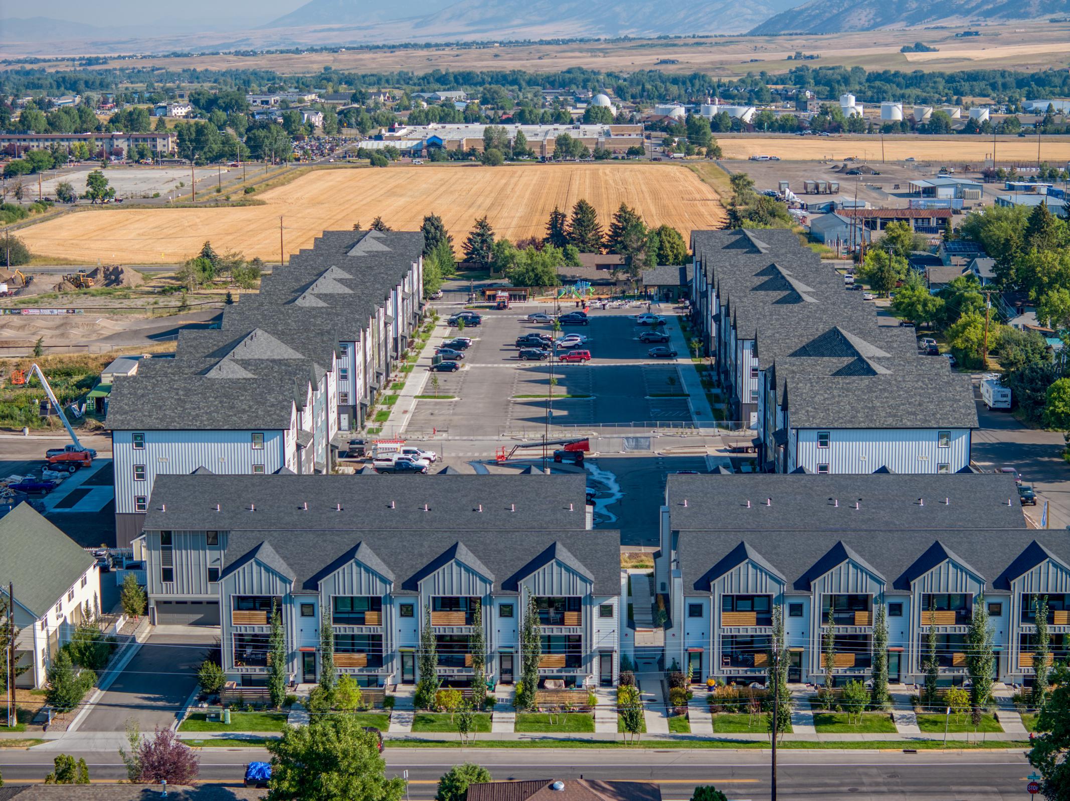 Aerial view of a residential area featuring two rows of multi-unit apartment buildings. Each building has a distinct roof design. The surrounding landscape includes fields, a parking lot filled with cars, and distant mountains under a clear blue sky. The scene showcases a blend of urban living and natural beauty.