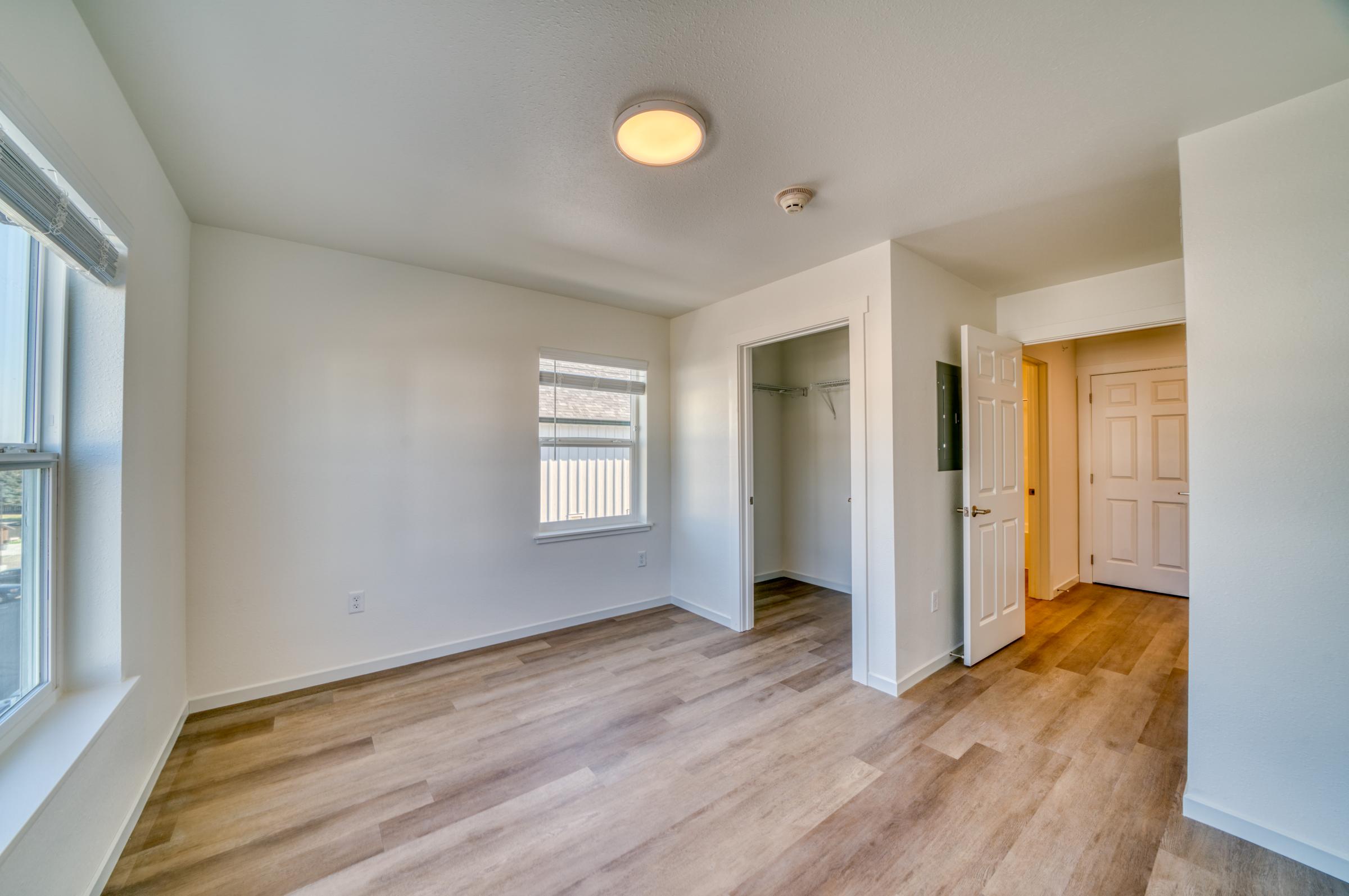 Bright, empty room featuring light-colored walls and wooden flooring. Two windows provide natural light, and there is a single ceiling light fixture. A doorway leads to a closet, with an adjacent hallway door visible. The space appears clean and newly finished, ideal for various uses.