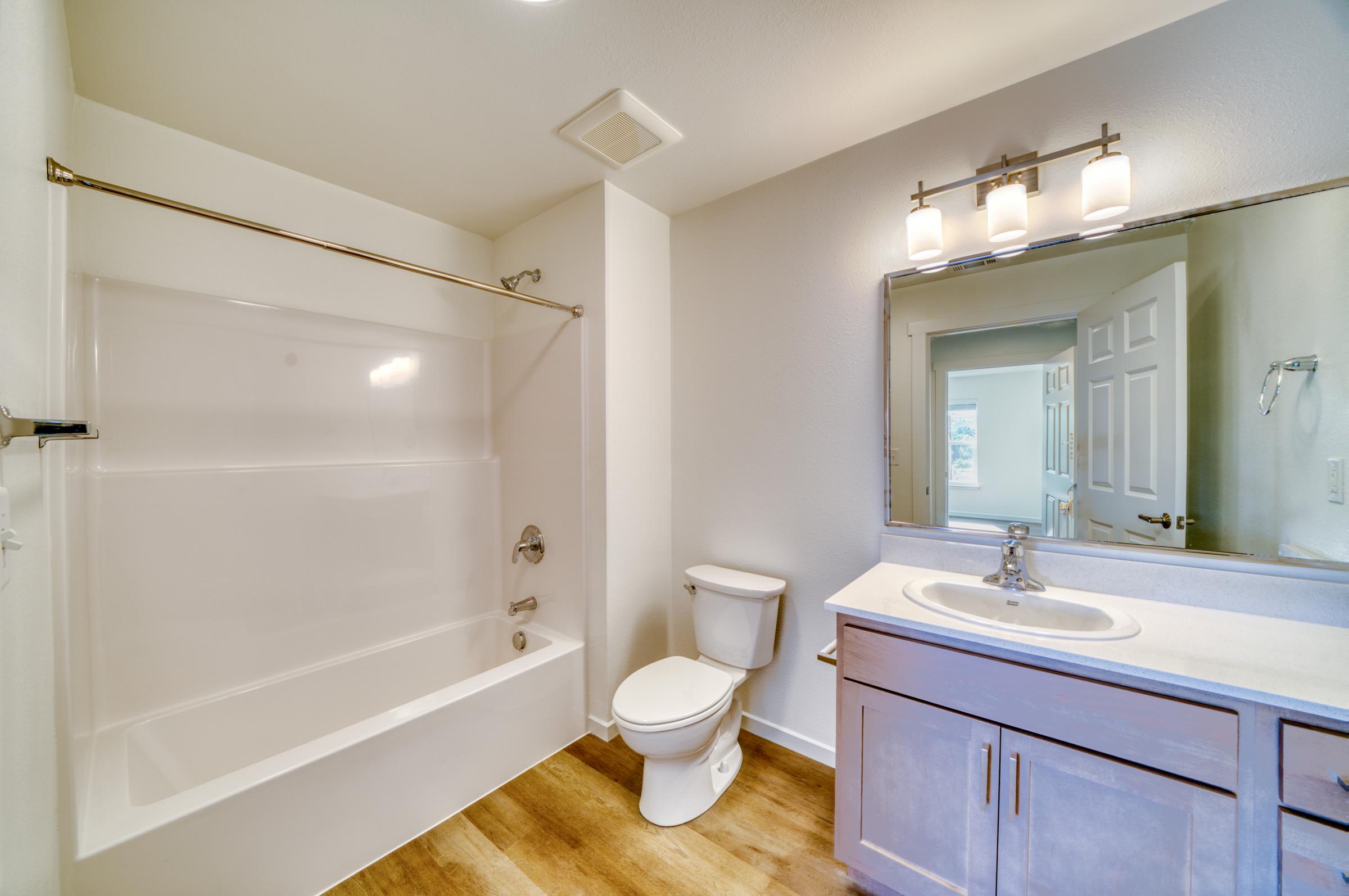 A clean and modern bathroom featuring a white bathtub with a shower curtain, a toilet, and a vanity sink with a mirror above it. The walls are painted in a light color, and there is wooden flooring. Natural light is visible through a door leading to another room.