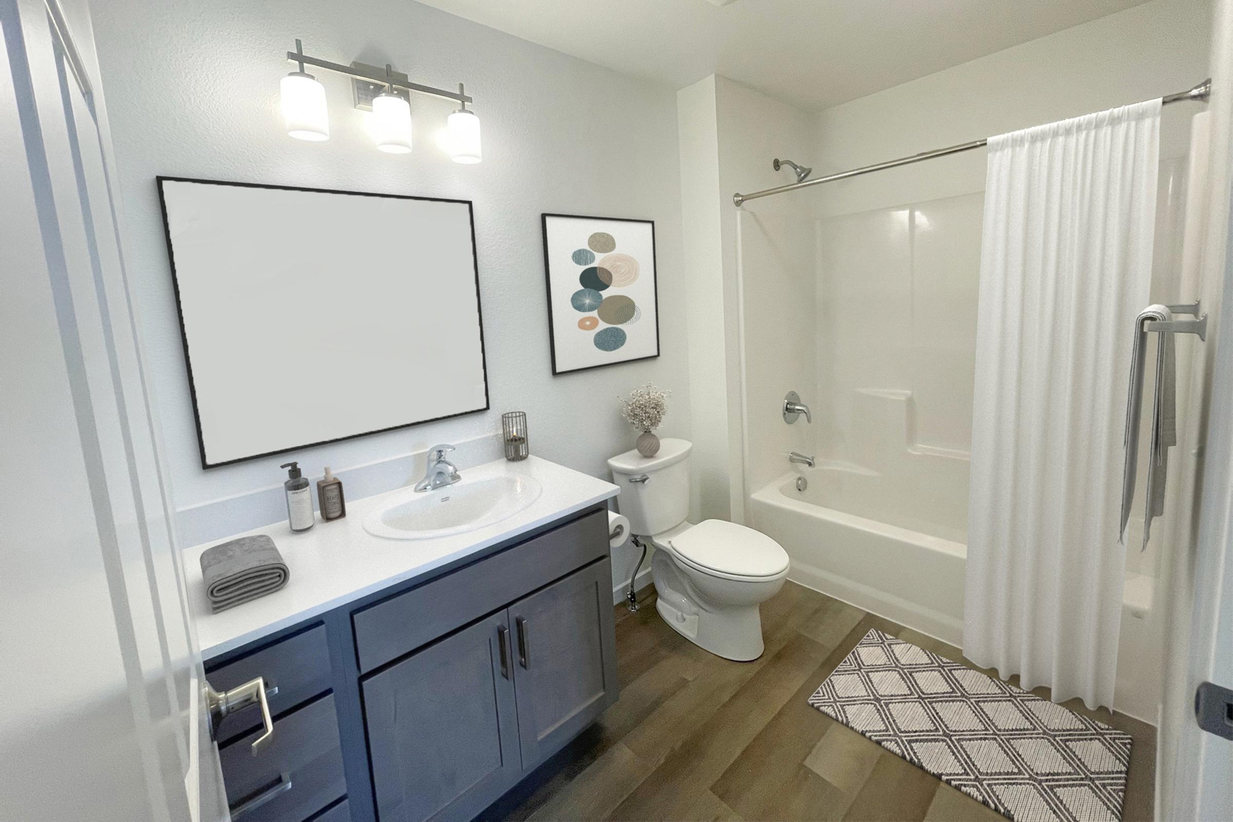 A modern bathroom featuring a white bathtub with a shower curtain, a white vanity with a round sink, and a large mirror above it. The walls are light gray, with a framed abstract art piece on one wall. The floor has wooden planks and a patterned bath mat, and there are toiletries neatly arranged on the counter.