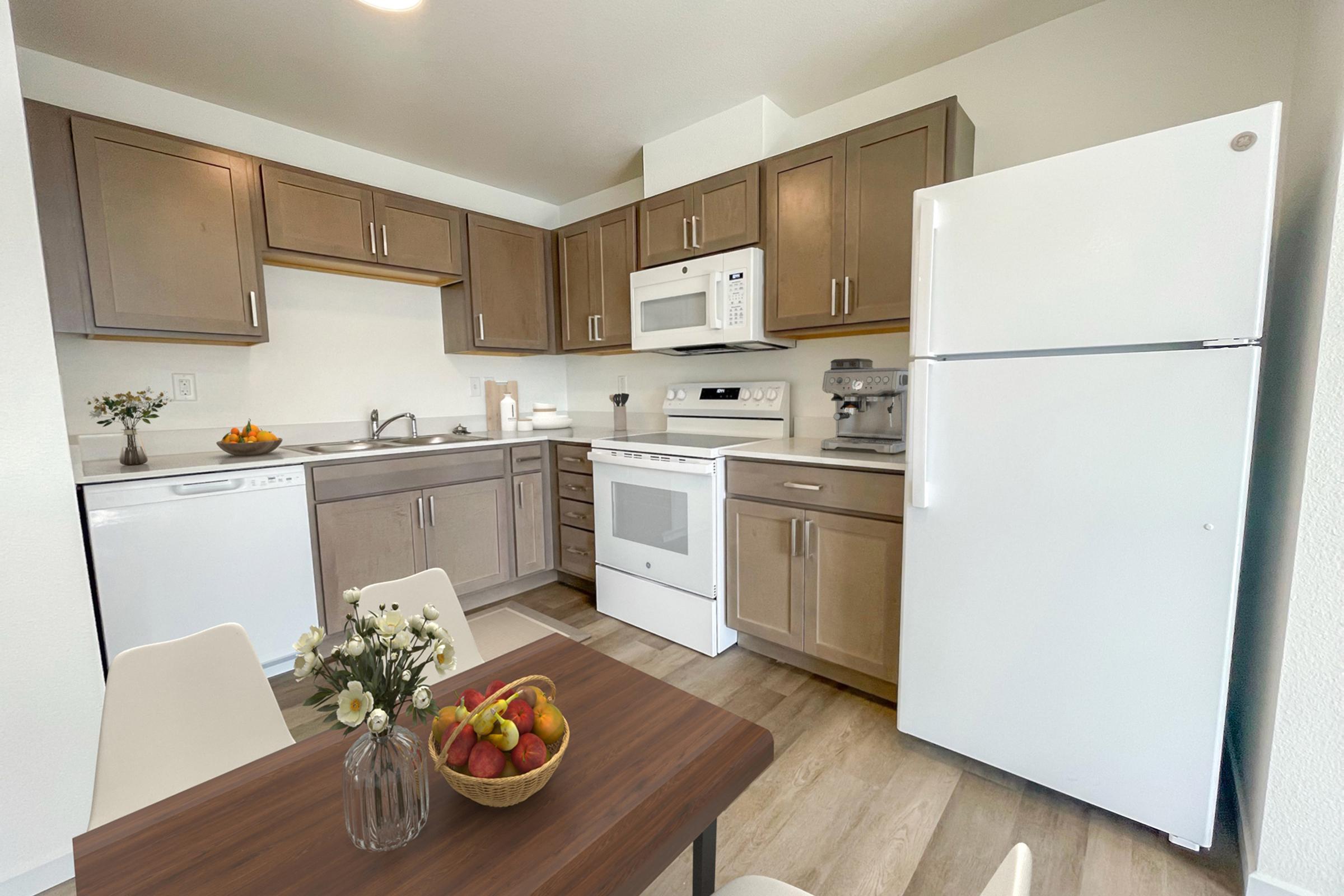 A modern kitchen featuring wooden cabinets, stainless steel appliances, and a white refrigerator. There is a dining table with a vase of flowers and a bowl of fruit, along with a sink, microwave, and oven, creating a cozy and inviting atmosphere. Natural light brightens the space.