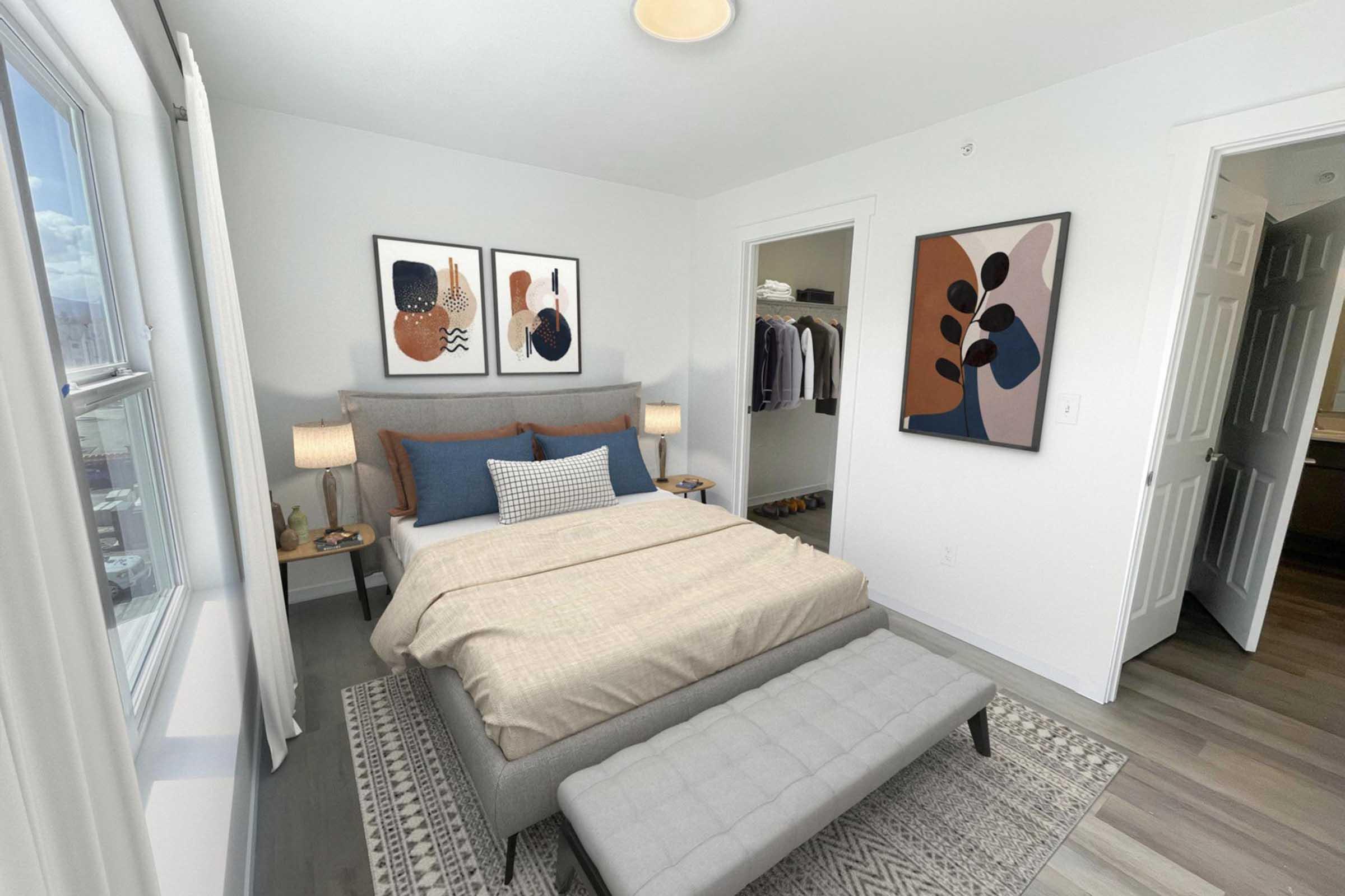 A modern bedroom featuring a neatly made bed with beige and blue bedding. There are two framed artworks on the wall, bedside tables with lamps, and a cozy bench at the foot of the bed. A closet is visible in the background, and the space has large windows allowing natural light.