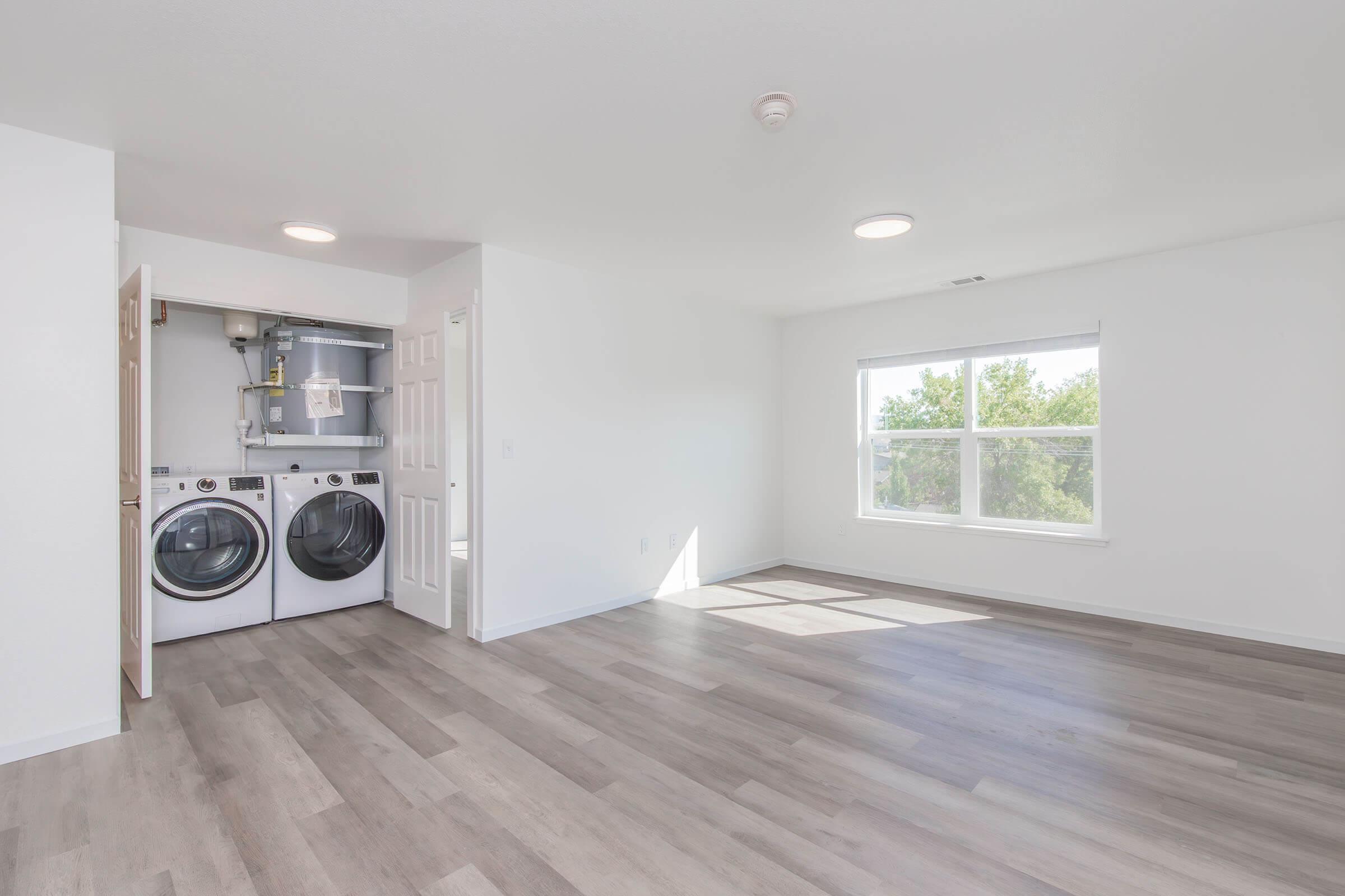 A bright and spacious room with light-colored walls and hardwood flooring. On one side, there is a laundry area with a washer and dryer. A large window allows natural light to flood the space, enhancing the open and airy atmosphere of the room.