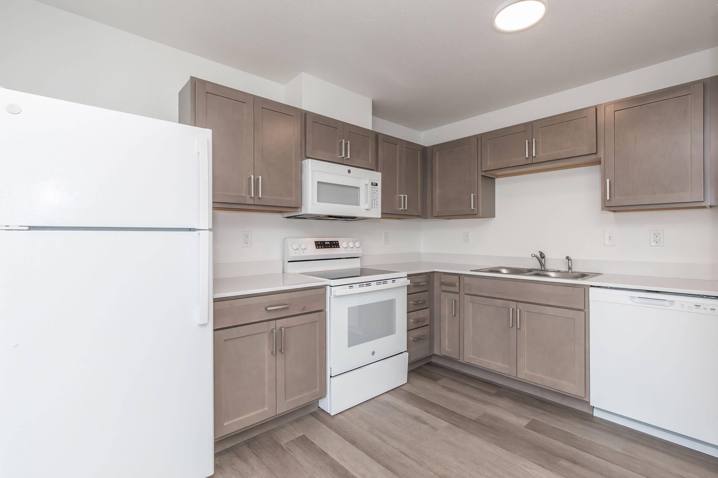 A modern kitchen featuring light gray cabinets, a white refrigerator, an oven with a stovetop, a built-in microwave, and a sink. The countertop is beige, and there is a white dishwasher. The flooring is a light wood finish, creating a bright and open space.