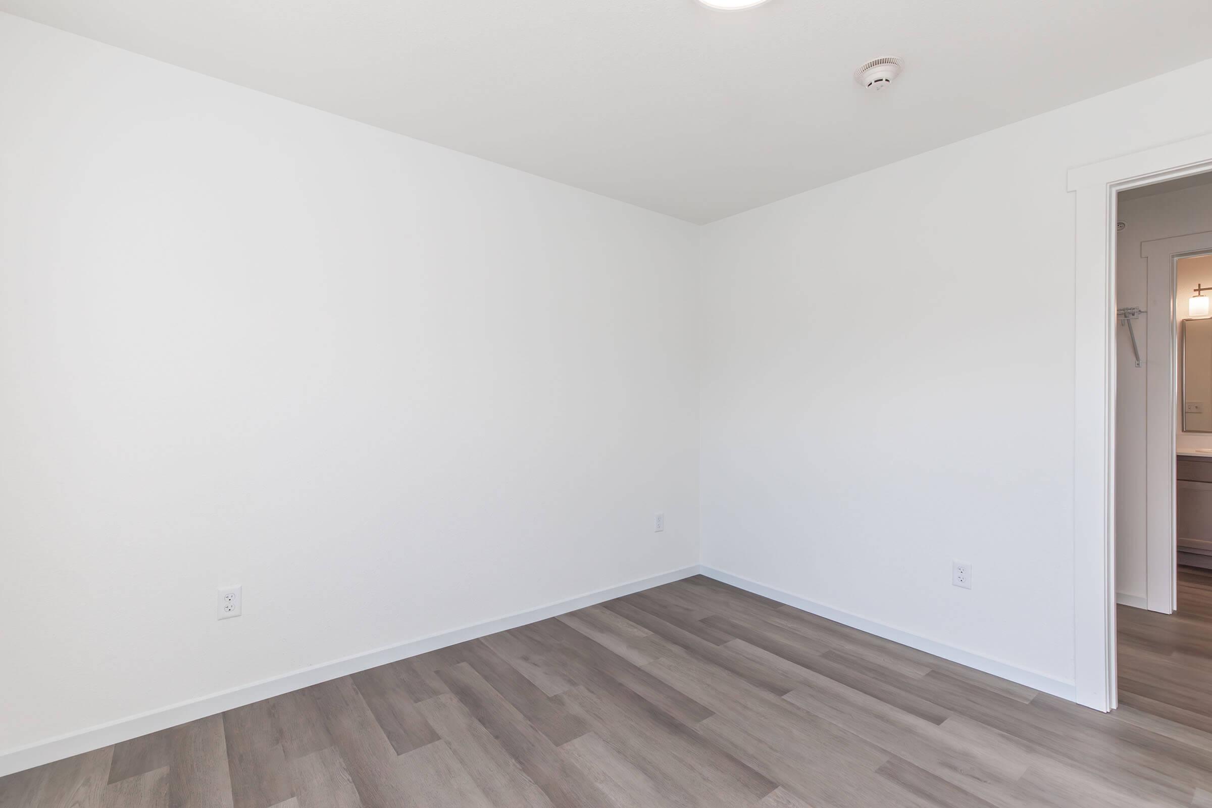 A bright, empty room with white walls and a light wood floor. There is a small ceiling light fixture, and a doorway leads to another area, hinting at additional space. The overall ambiance is clean and minimalist, suitable for various purposes.