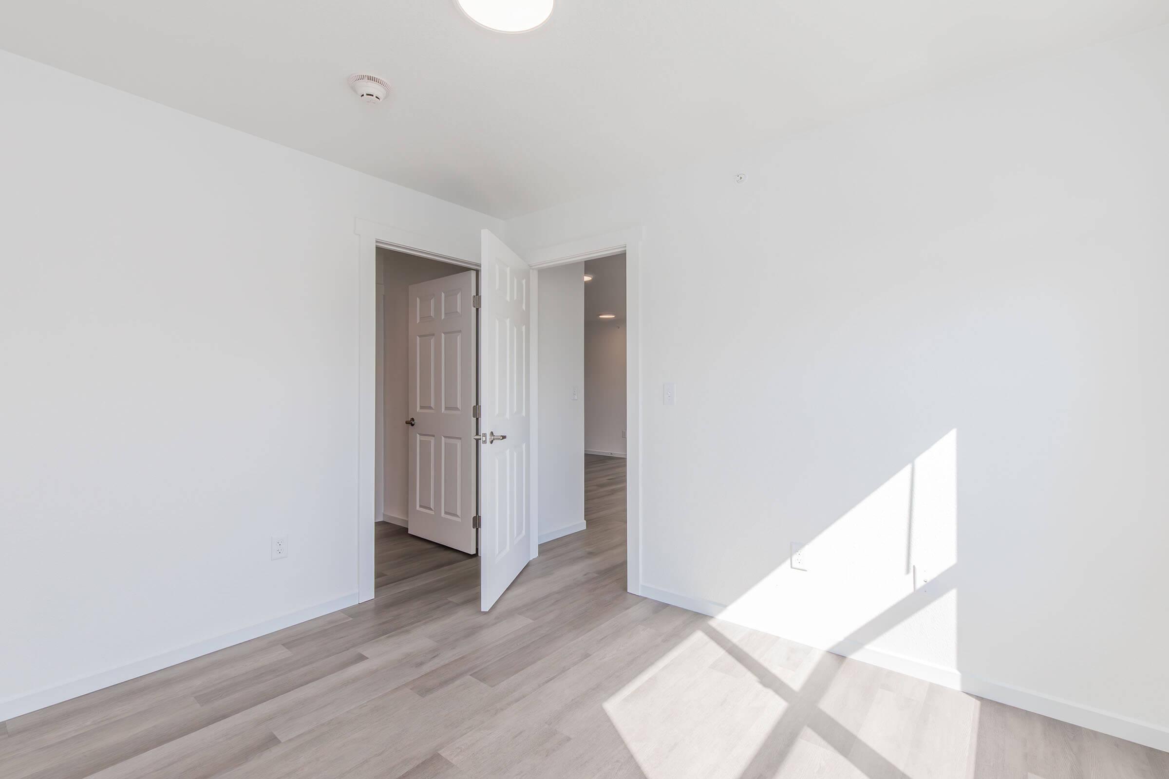 A bright, empty room with white walls and light-colored laminate flooring. A door is partially open, leading to another room. Natural light streams in from a window, creating shadows on the floor. The overall atmosphere is clean and modern.