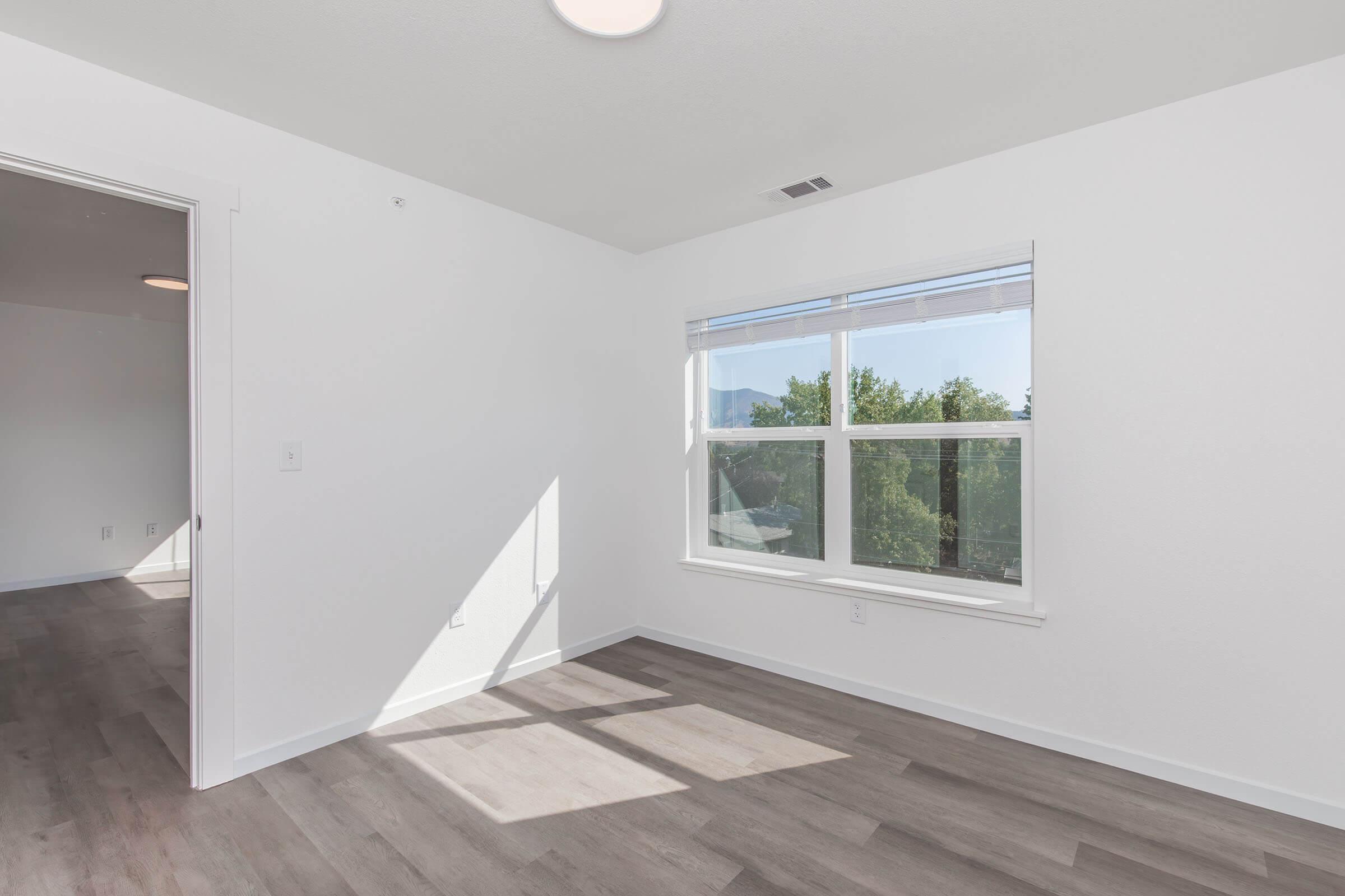 A bright, empty room with light-colored walls and a window that allows natural light to flow in. The floor is a light hardwood, and there is a glimpse of greenery outside. The room features a simple modern design with no furniture, highlighting its spaciousness.