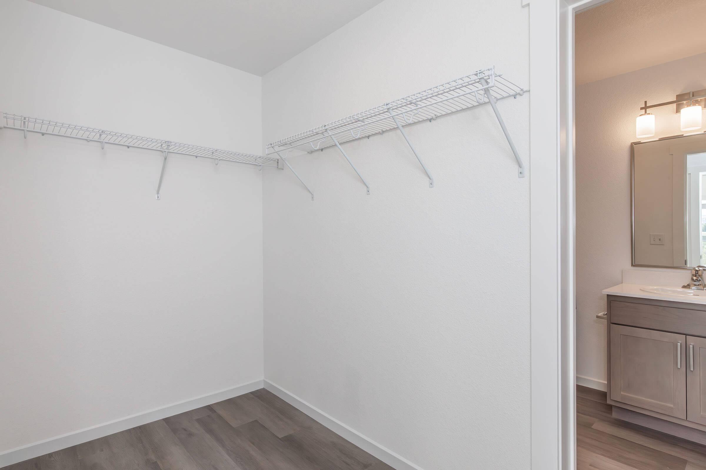 Empty walk-in closet with white walls and wood-like flooring. Features two wire shelves on opposite walls. A doorway on the right leads to a bathroom with a light fixture and sink visible.