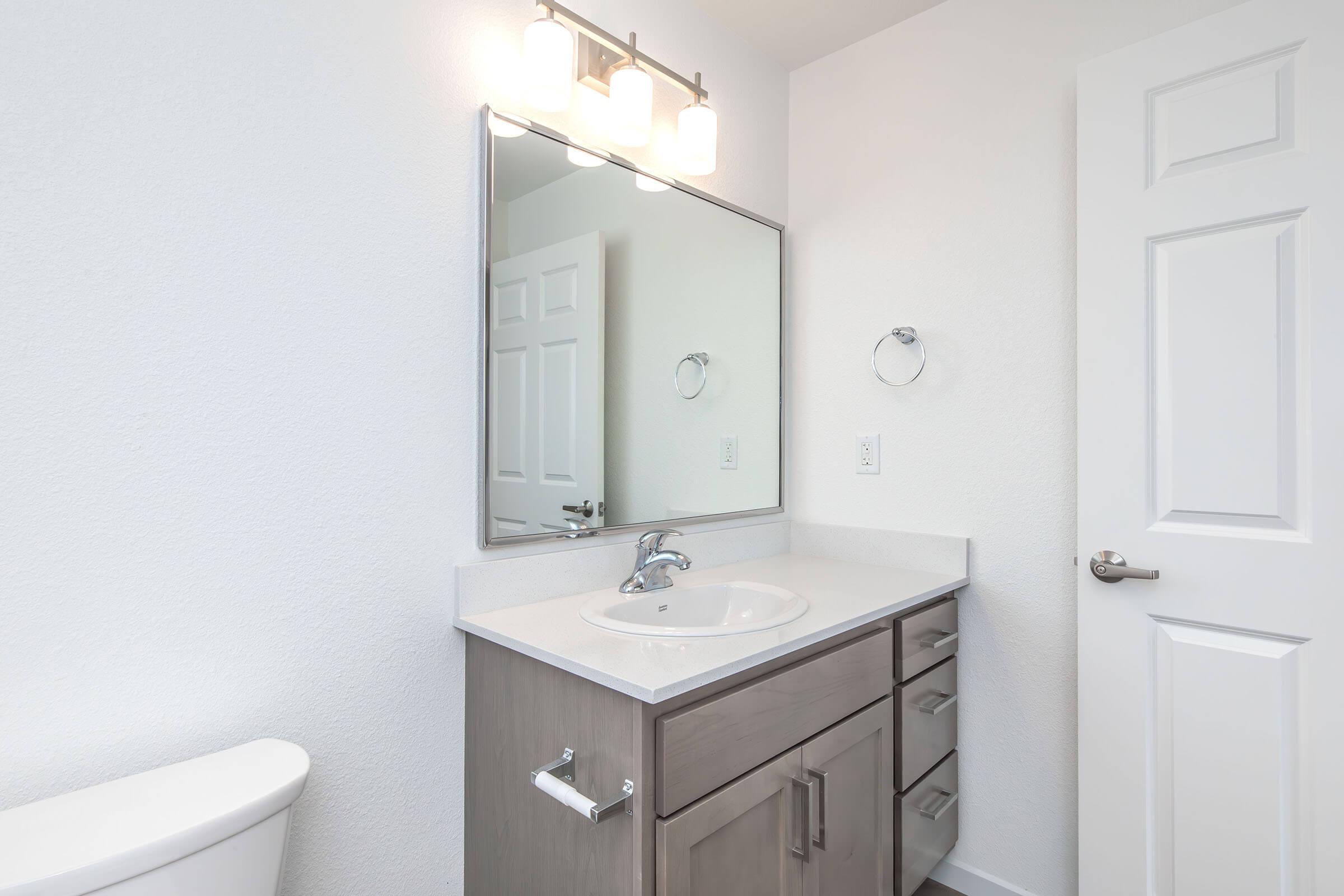 A modern bathroom featuring a sink with a countertop, a large mirror, and three light fixtures above. There are drawers and a towel holder beside the sink. A white toilet is visible on the left side, and the walls are painted a light color, creating a clean and spacious ambiance.