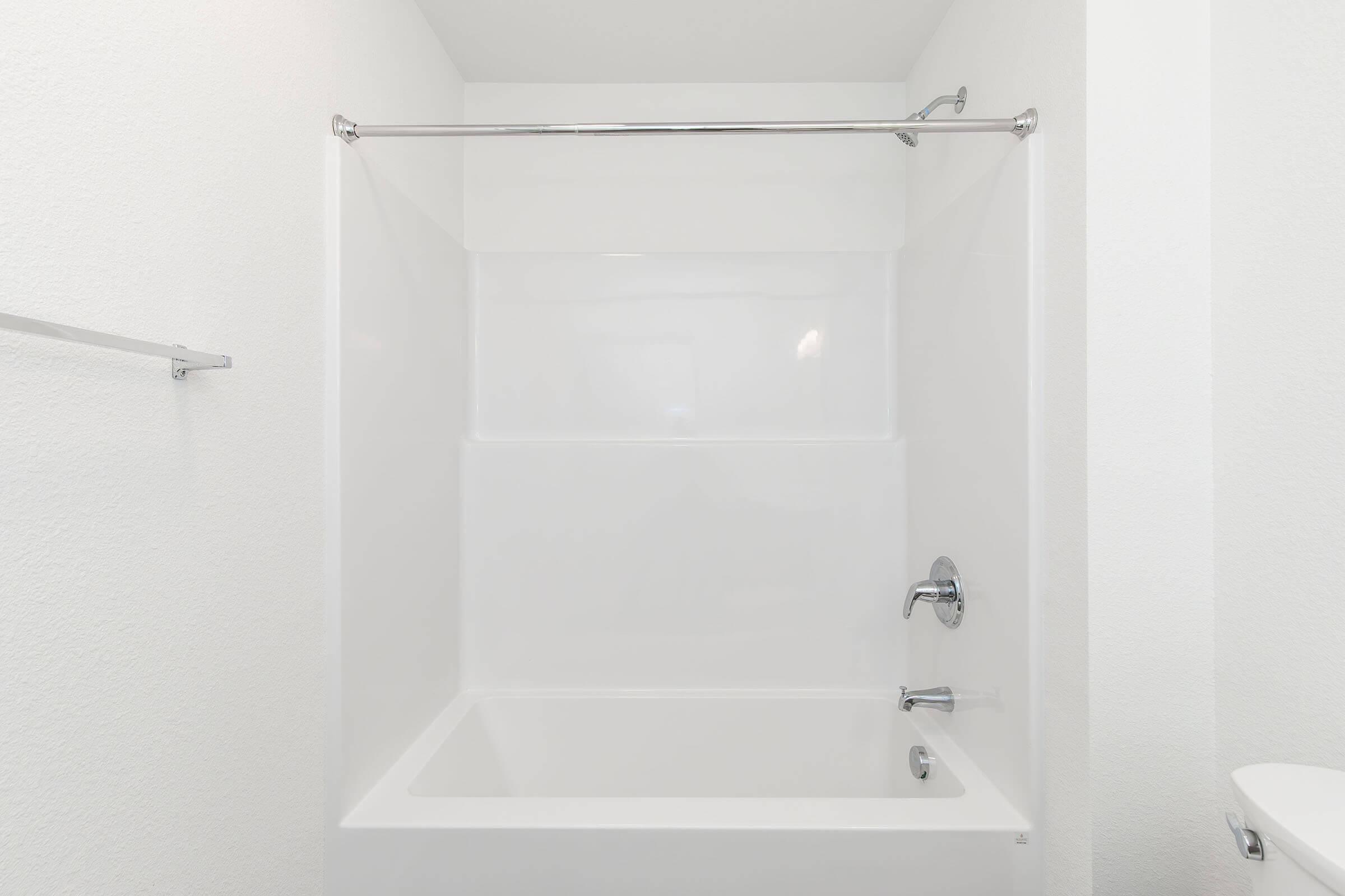 A clean and modern bathroom featuring a white tub and shower area. The walls and tub are smooth and white, with a chrome showerhead and faucet. A shower curtain rod can be seen, offering functionality to the space. The overall look is minimalist and bright, emphasizing a fresh and inviting atmosphere.